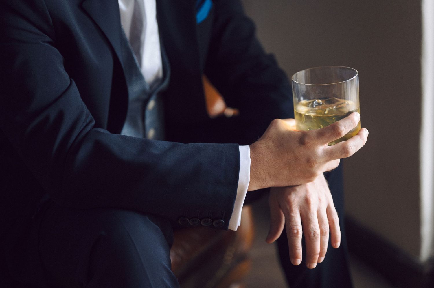 Person in navy suit holding glass of whiskey or bourbon while sitting in contemplative pose.