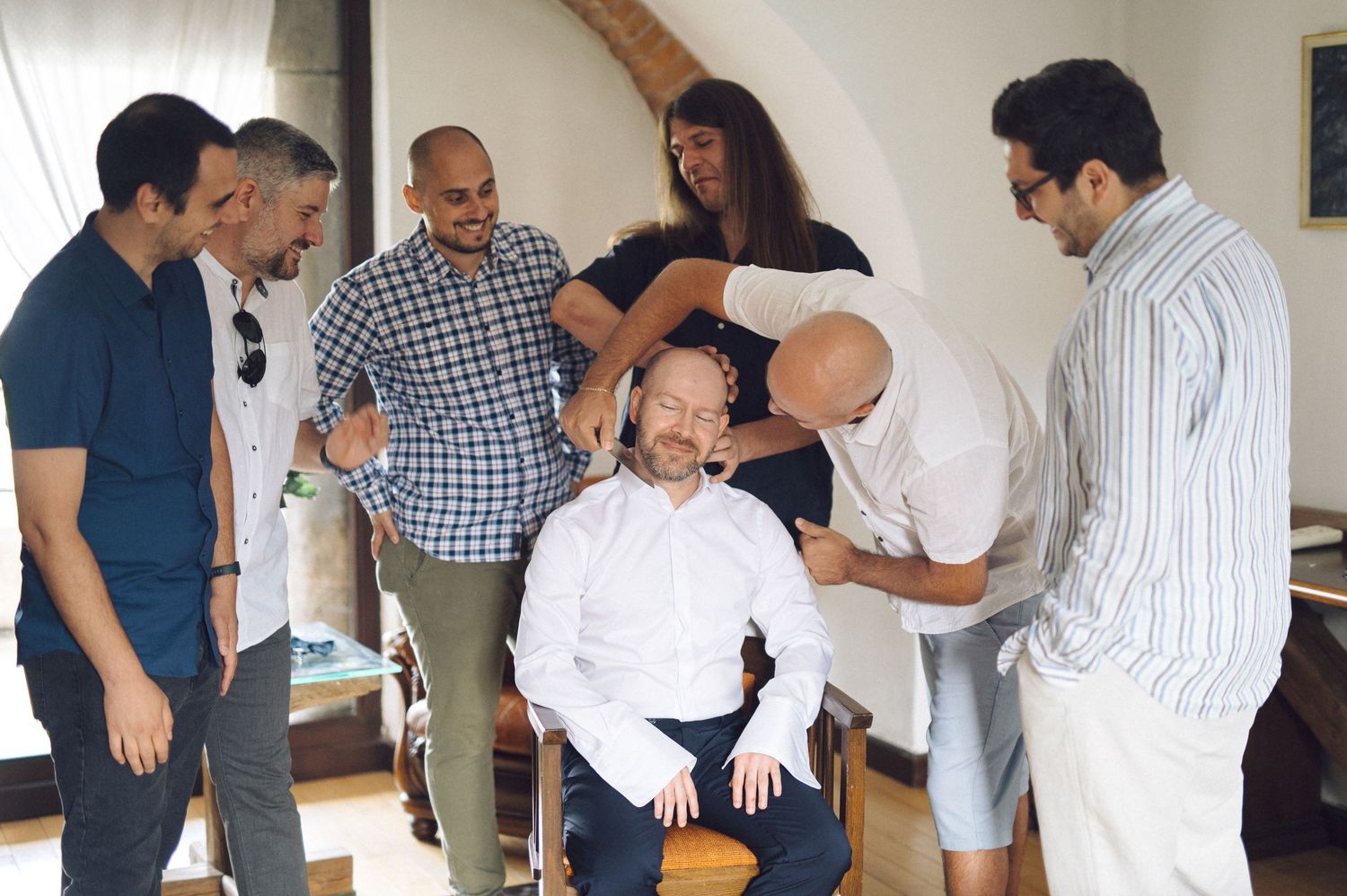 A group of people gather around someone seated in a chair during what appears to be a grooming or styling session.