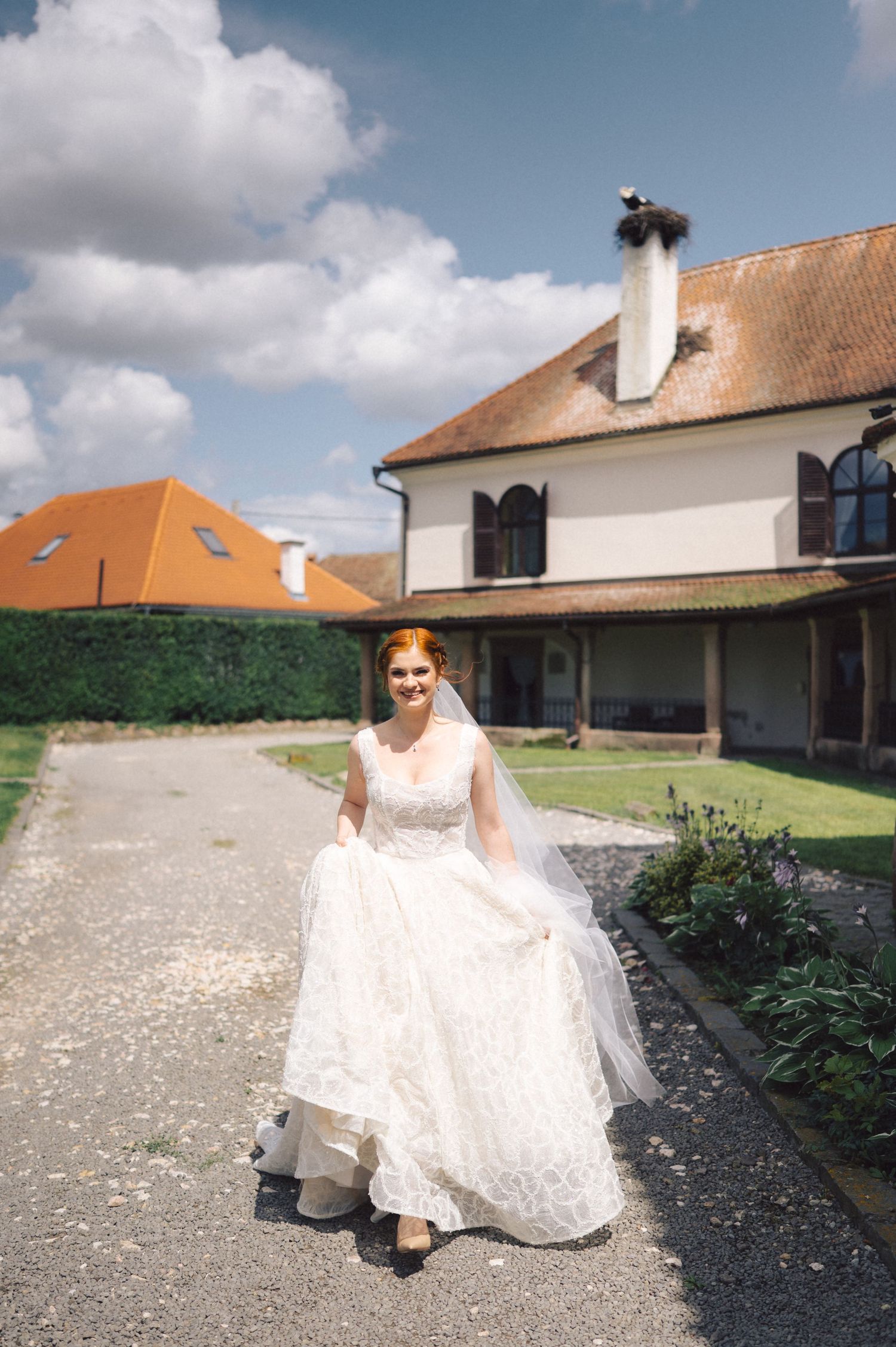 A bride in a lace white wedding gown stands on a gravel driveway in front of a white country house with an orange roof.
