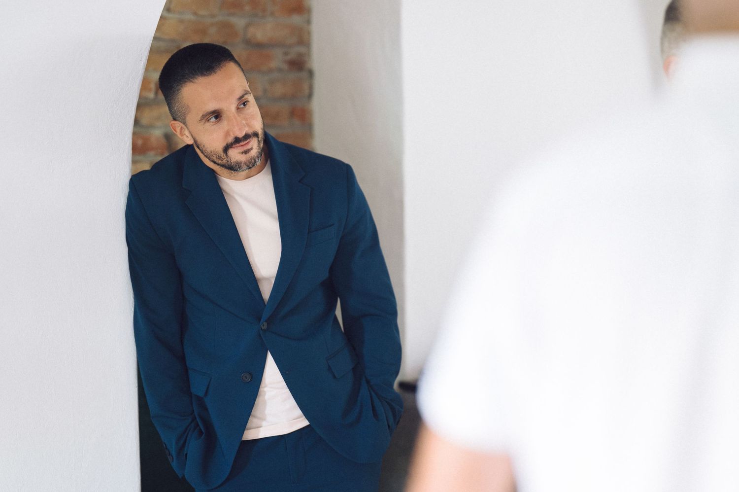 Professional portrait of someone in a navy blue suit jacket and white shirt leaning against a light colored wall.