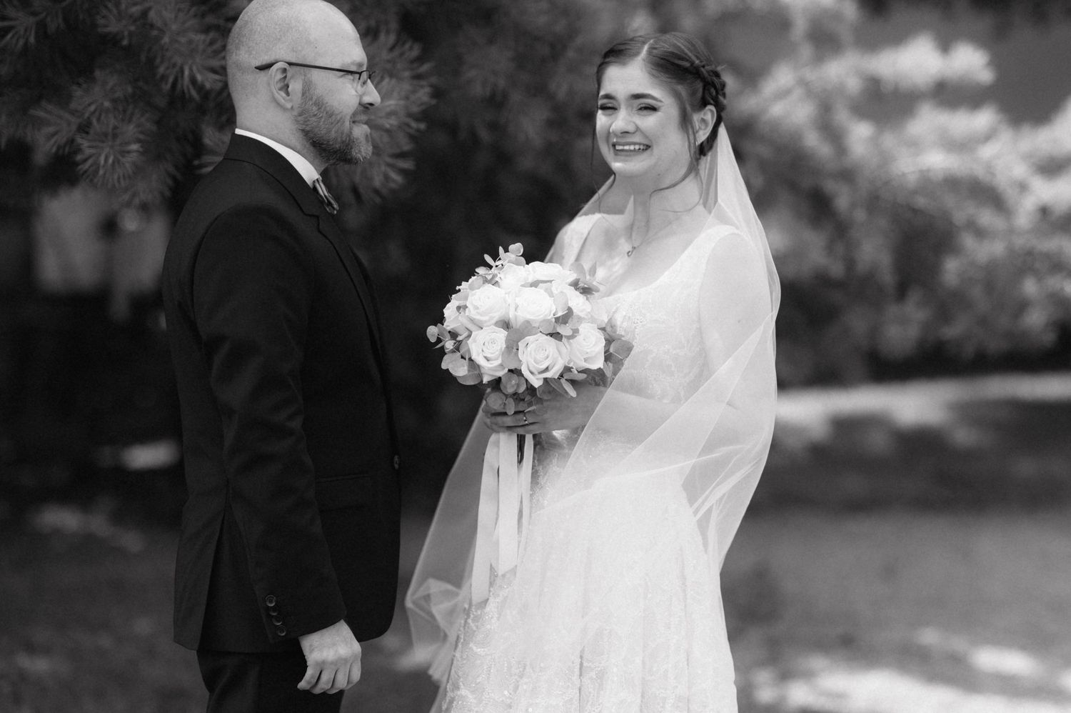 A black and white wedding photo shows a couple in a romantic outdoor setting with the bride holding a white floral bouquet.