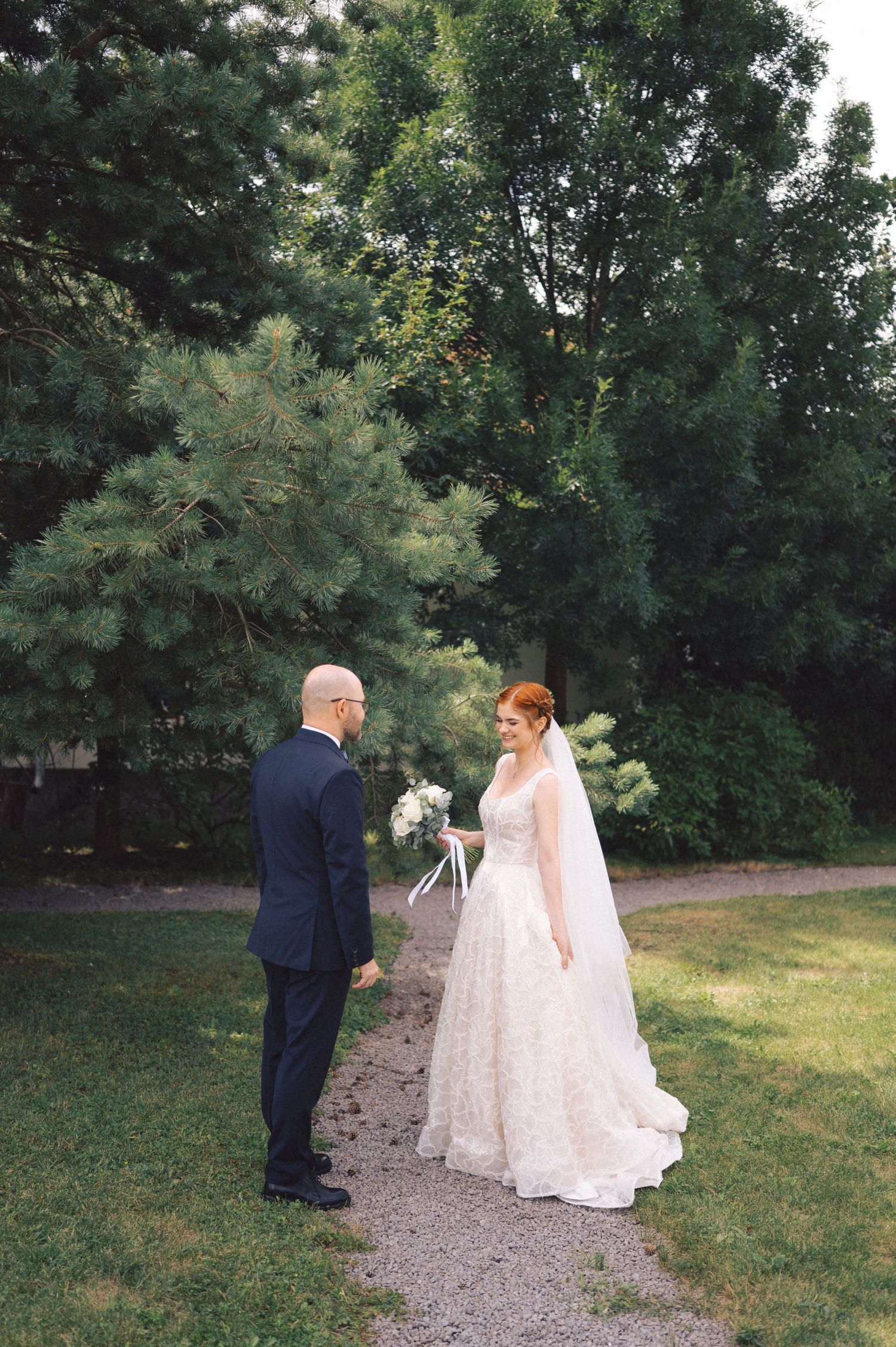 Wedding couple walks together on a gravel path surrounded by lush green trees on a sunny summer day.