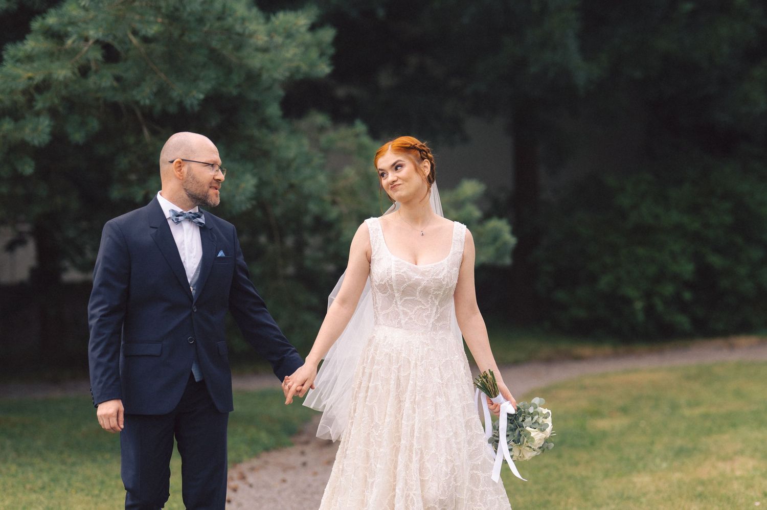 A couple walks together on a path outdoors, the person on left wears navy suit while right wears light pink wedding dress.