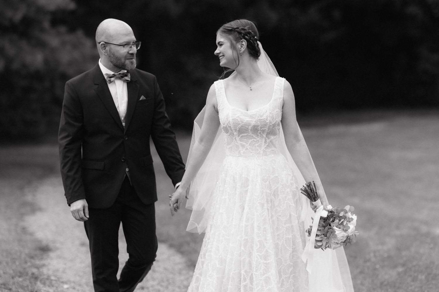 A bride and groom walk hand in hand on their wedding day in a black and white outdoor photograph.