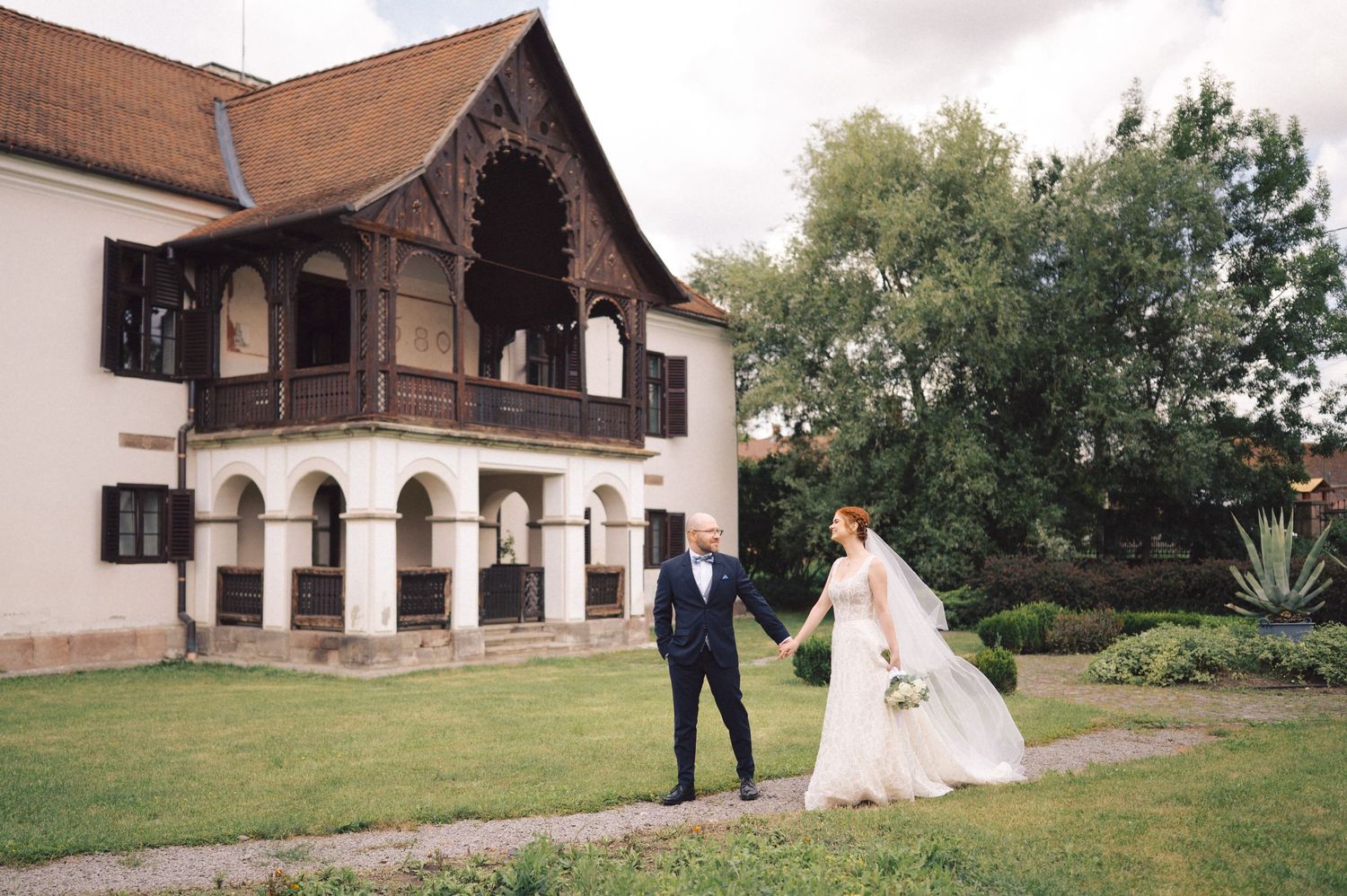 Wedding party celebrates outdoors at rustic venue with traditional buildings and green lawns in background.