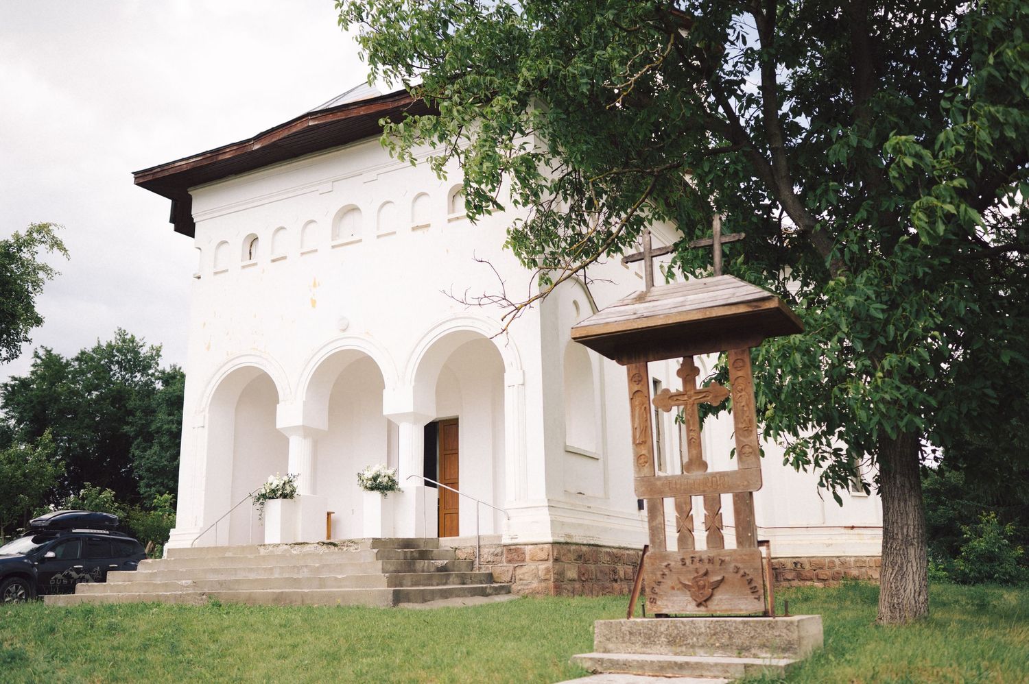 White church building with arched entranceway and wooden bell tower standing beside a tree in rural setting.