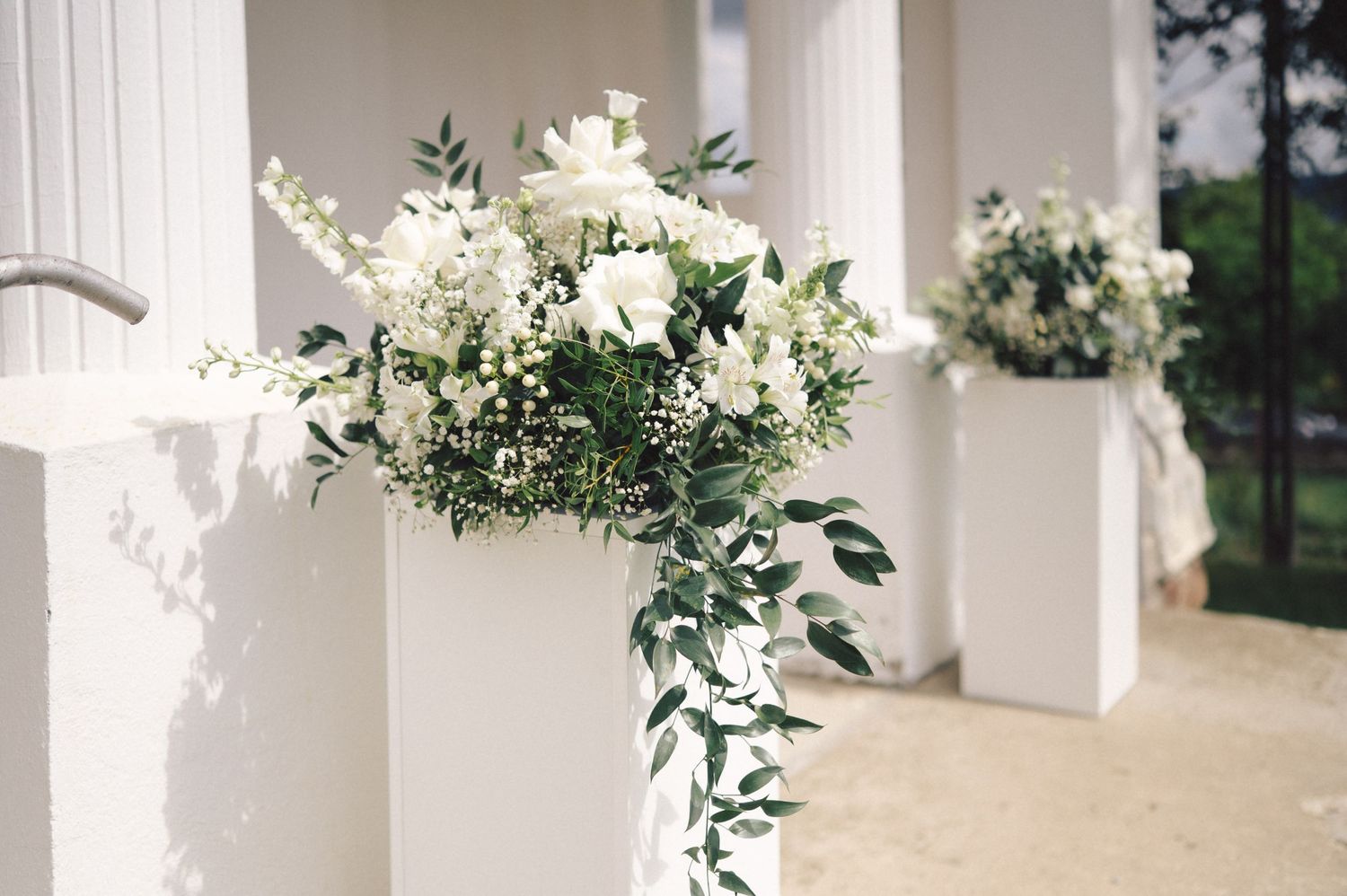 Elegant white floral arrangement with cascading greenery displayed on a classic white column pillar for a wedding ceremony.