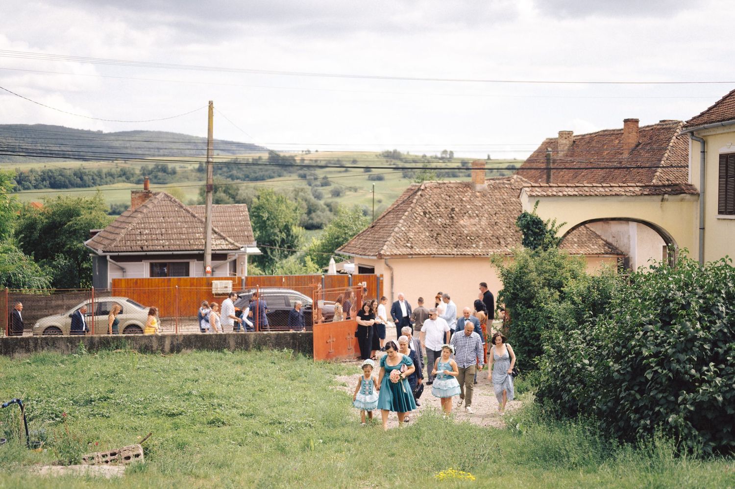 A group of people gather at a rural site with historic buildings and green rolling hills in the background.