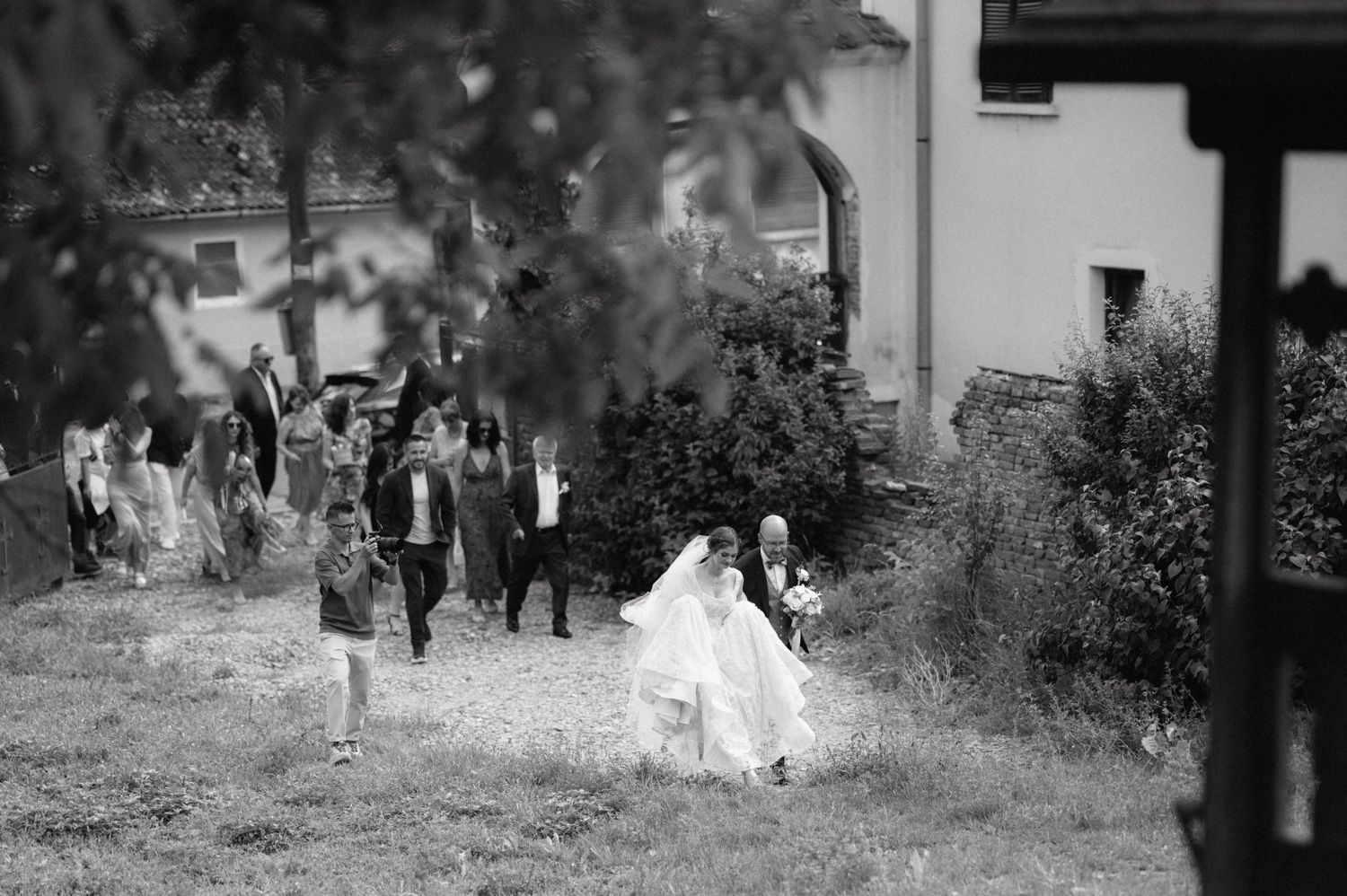 A wedding party walks along an outdoor path in black and white, with the bride in white leading the procession.