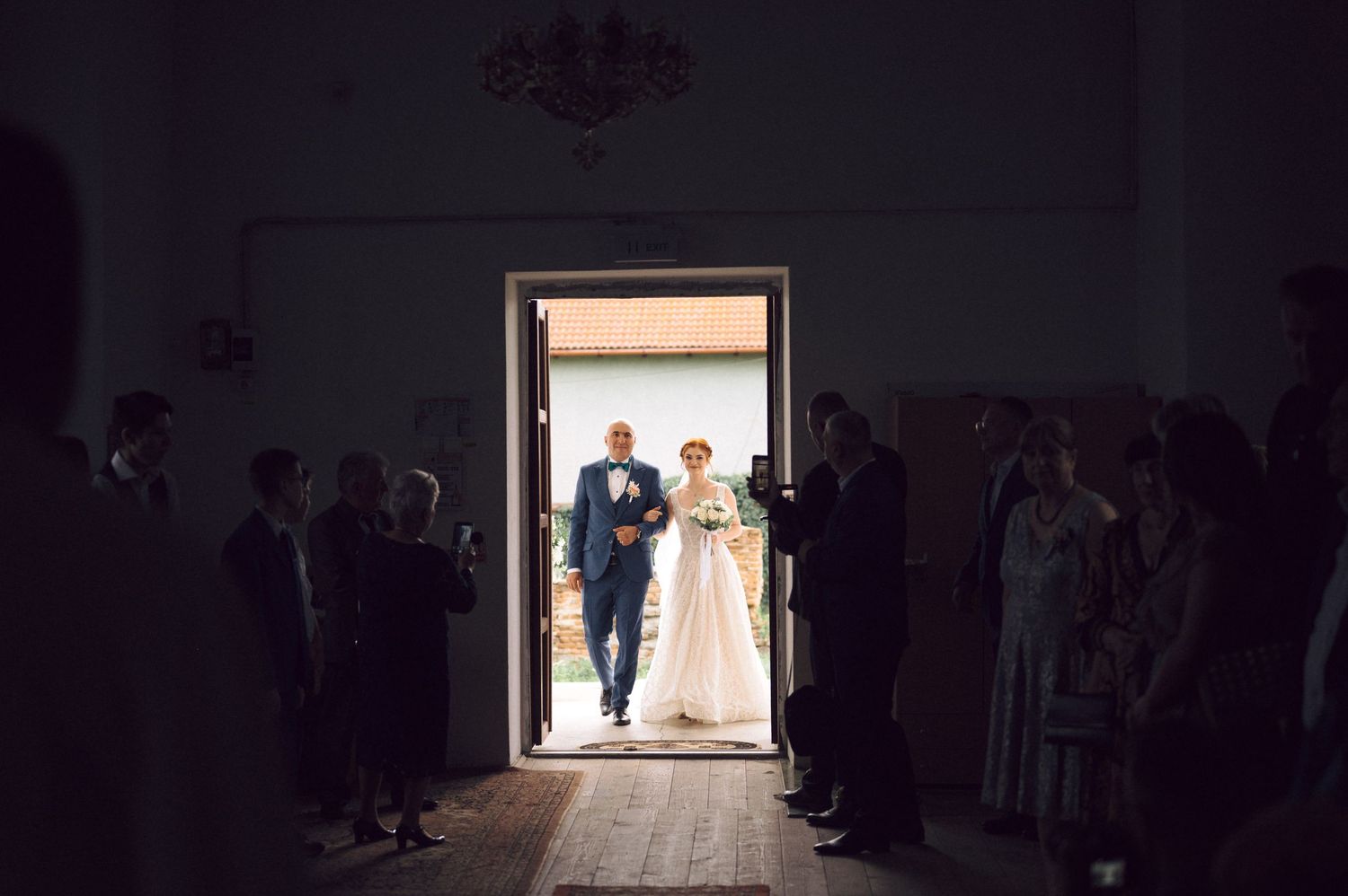 Silhouetted wedding guests line the walls of a dark room as light streams through a doorway at the entrance.