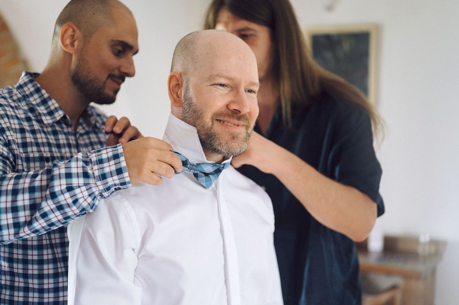 People help adjust a blue bow tie on someone wearing a white dress shirt at what appears to be a formal occasion.