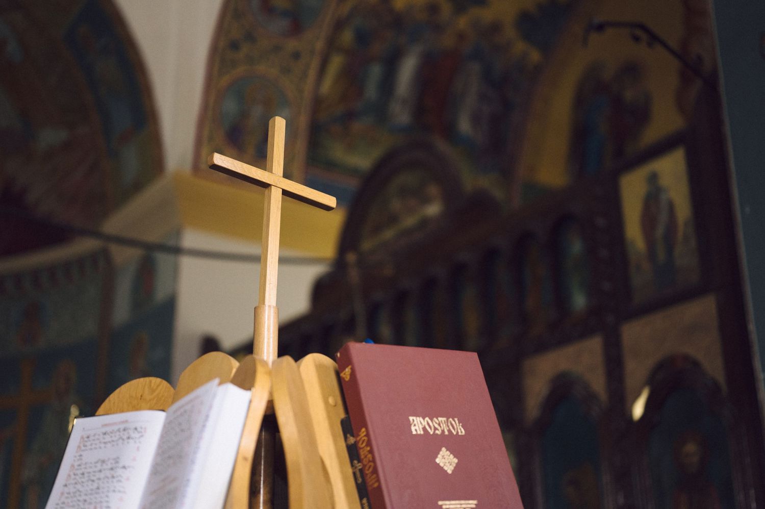 Church interior showing rows of wooden pews, religious books, and a golden cross illuminated in warm light.