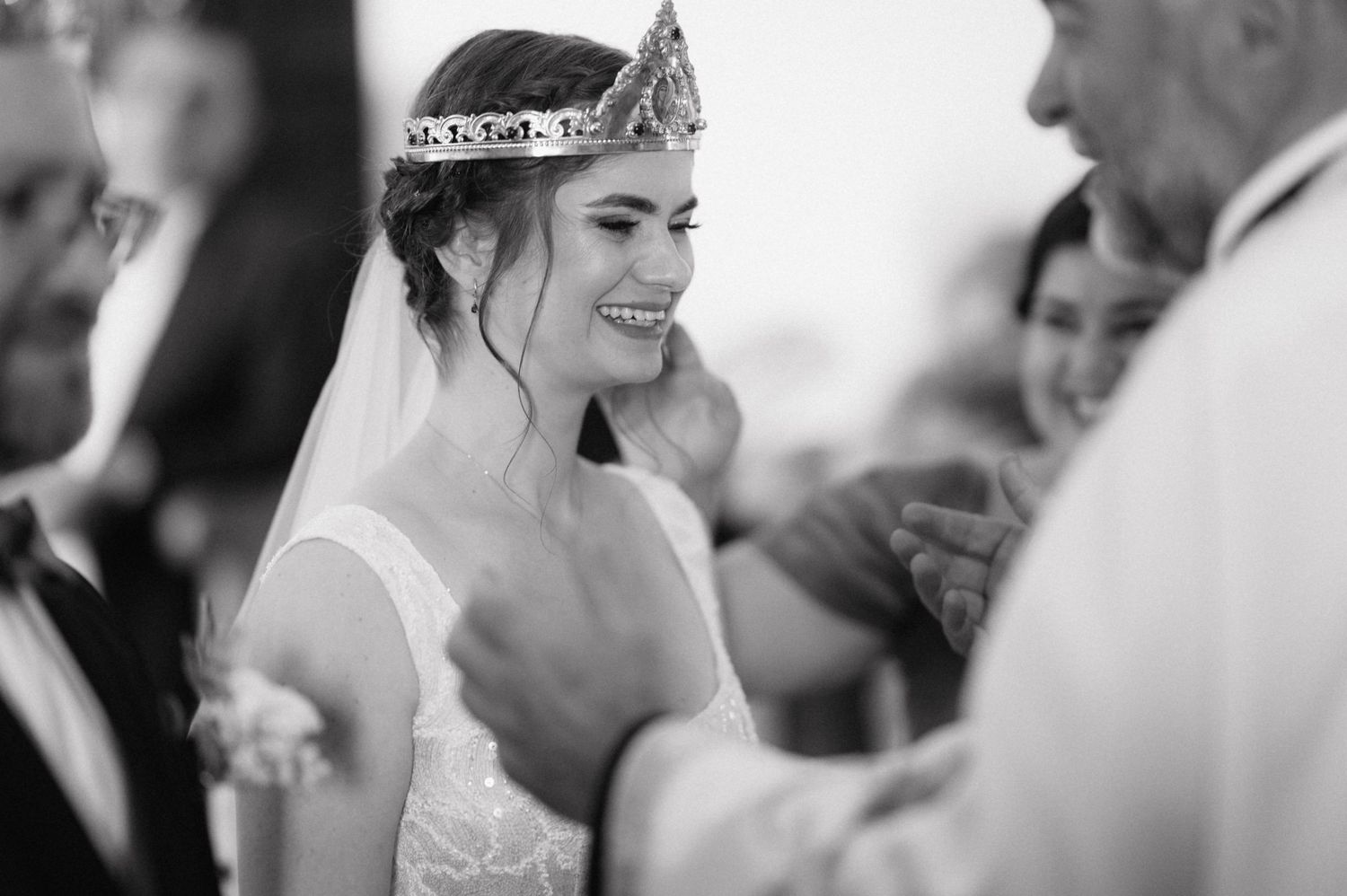 A joyful bride wearing a sparkling tiara and white dress smiles during what appears to be a wedding ceremony.
