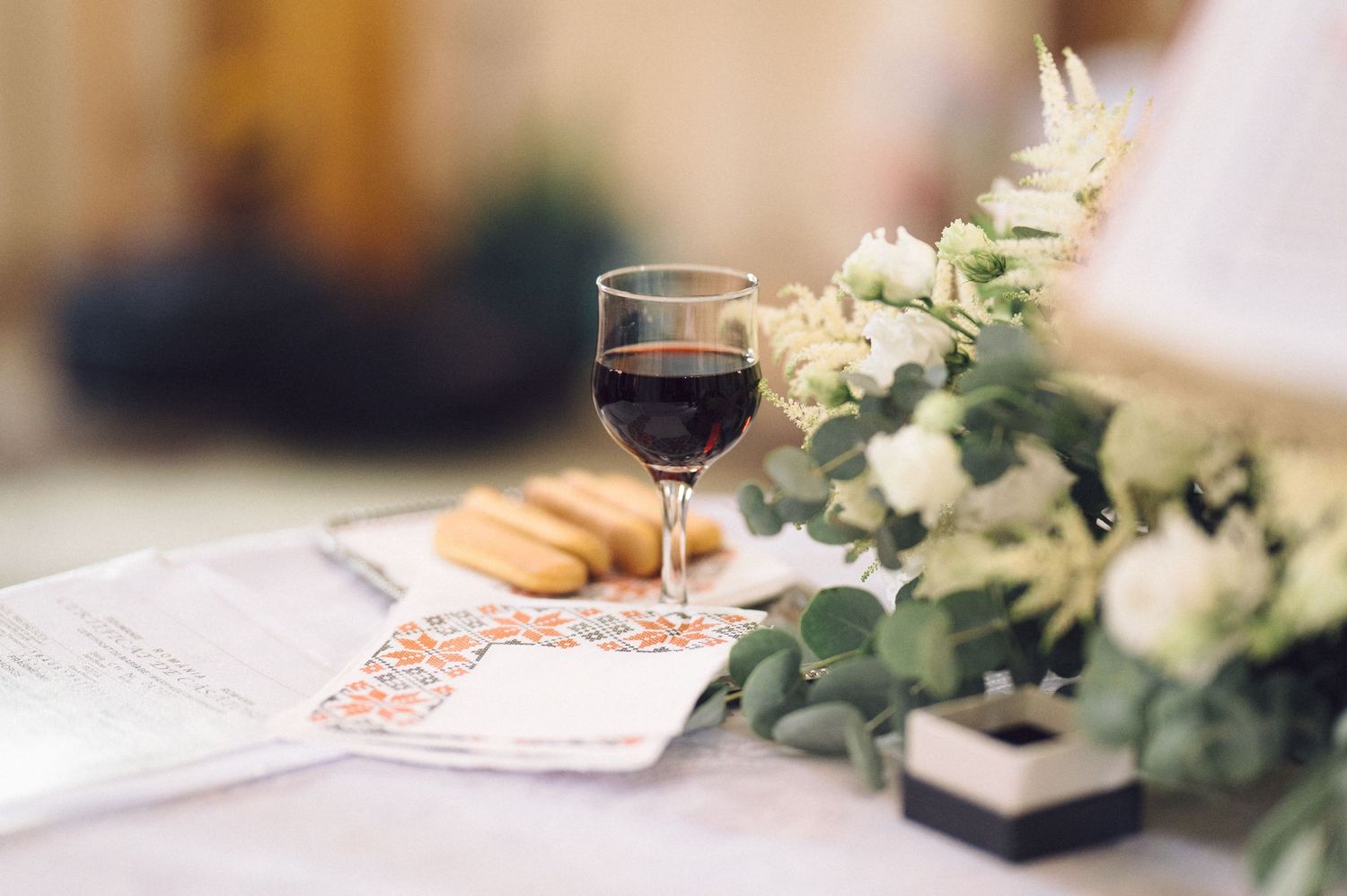 A glass of red wine sits beside white flowers and a newspaper on a table setting.