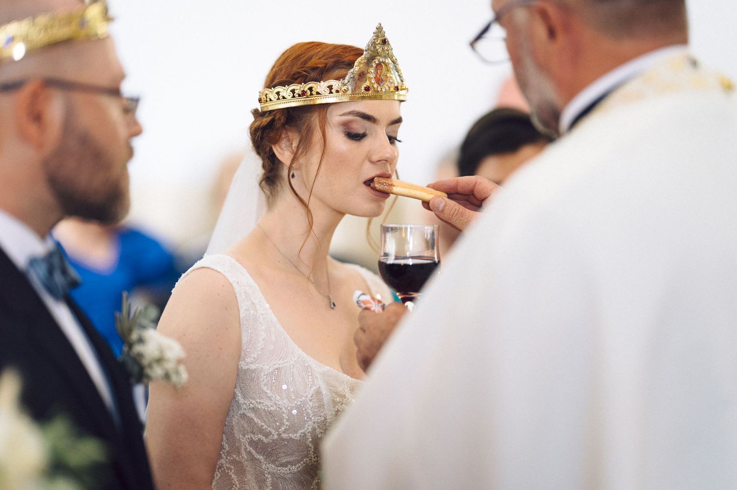 A bride wearing a golden crown and lace wedding dress sips from a wine glass during a wedding ceremony.