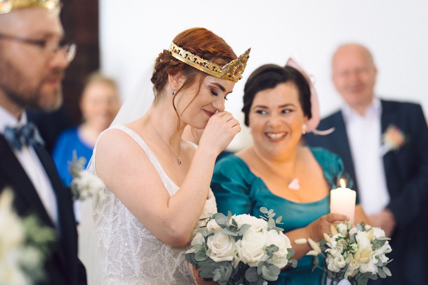 An emotional bride wearing a lace wedding dress and tiara holds a white rose bouquet during the ceremony.