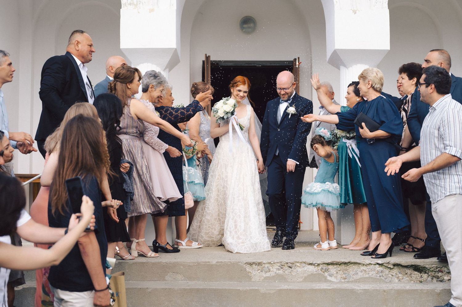 Wedding attendees stand on church steps tossing rose petals in a joyful celebration after ceremony.