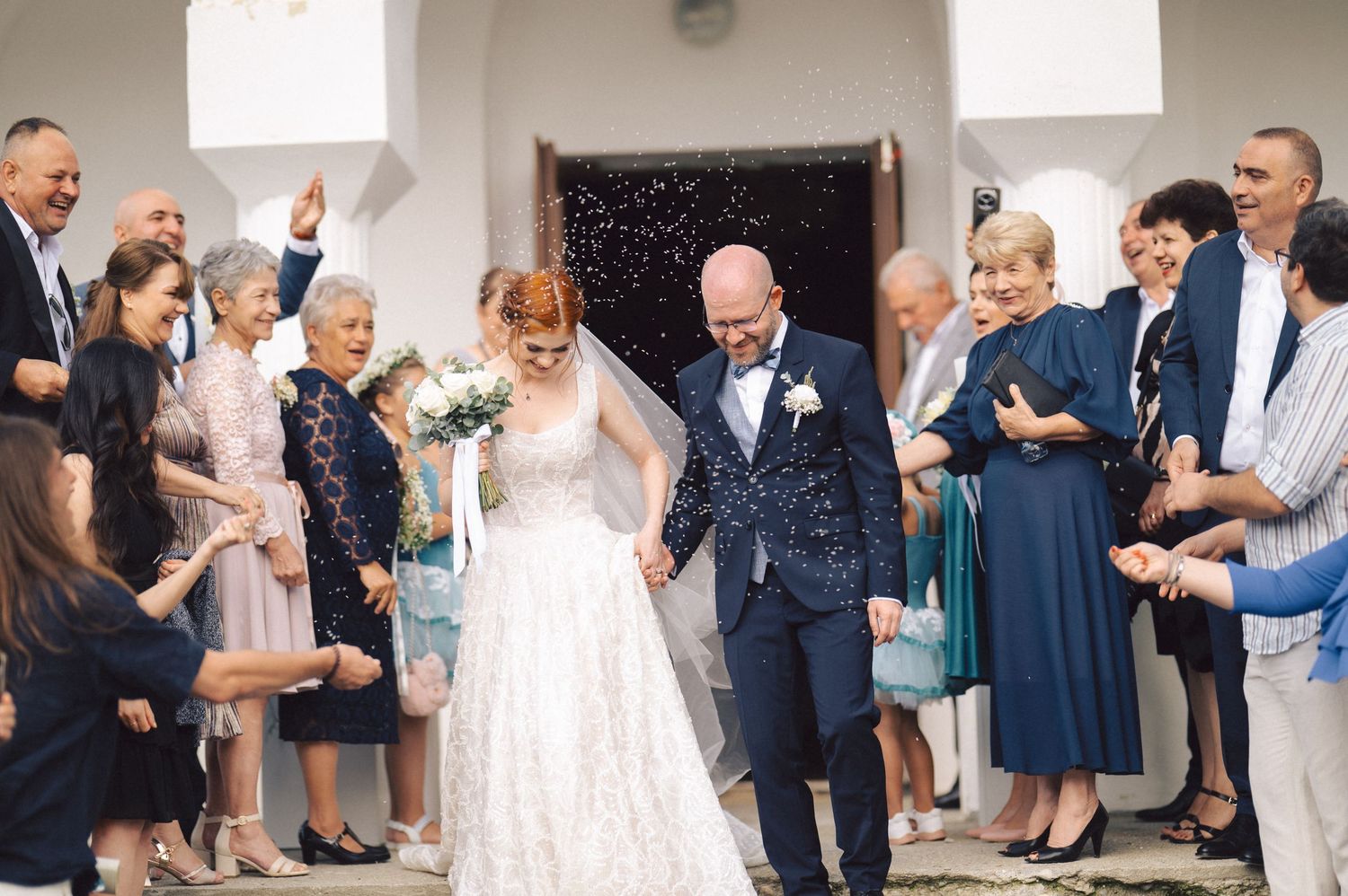 Wedding guests stand together laughing and smiling during an outdoor celebration with confetti falling in the air.