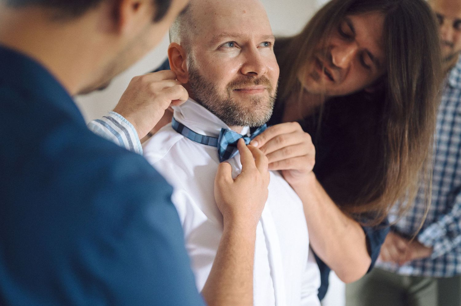 Someone helps adjust a light blue bowtie on a person wearing a white dress shirt while another person looks on and smiles.