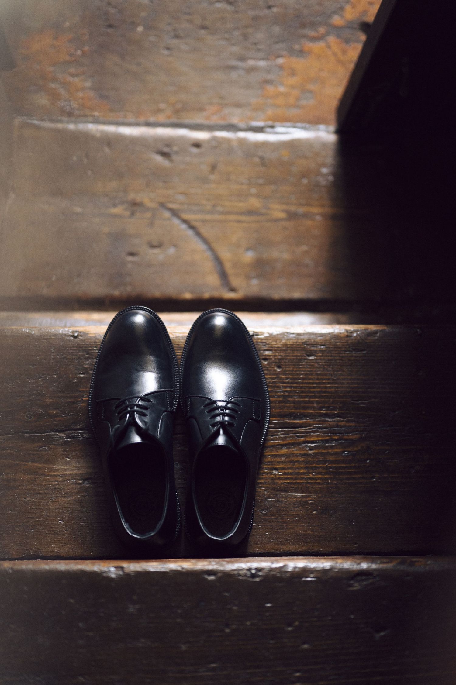 A pair of elegant black leather dress shoes with tassels resting on dark wooden stairs in moody lighting.