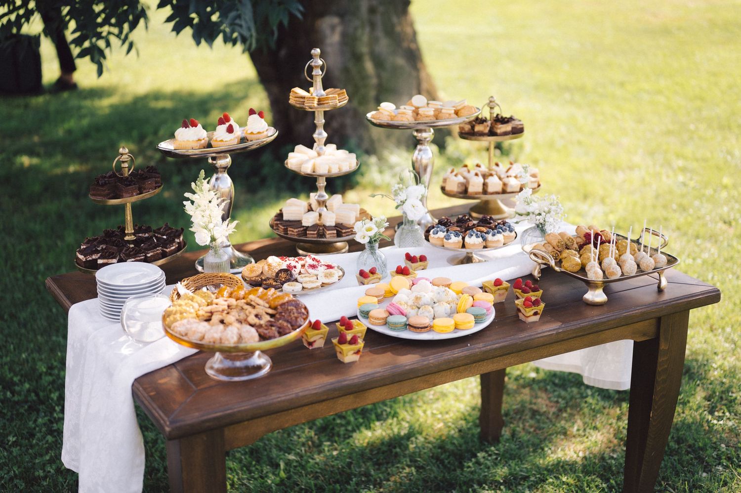 A rustic wooden table outdoors displays a beautiful array of cupcakes and desserts on tiered stands for a garden party.