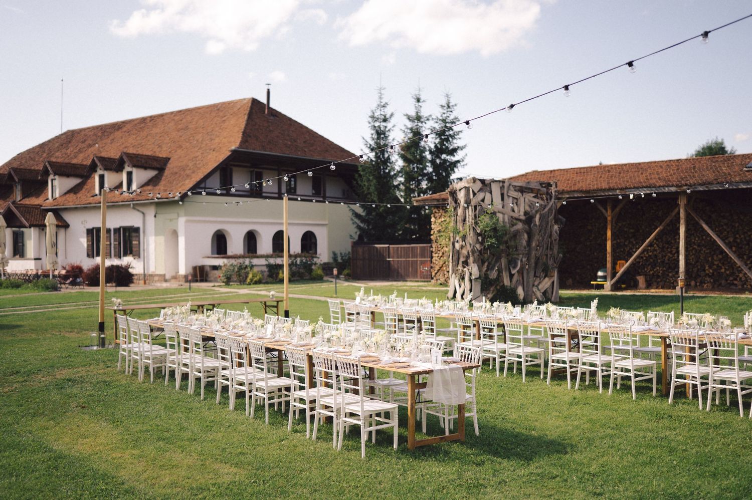 Elegant outdoor wedding setup with rows of white chairs and tables on a lawn in front of a large European-style villa.