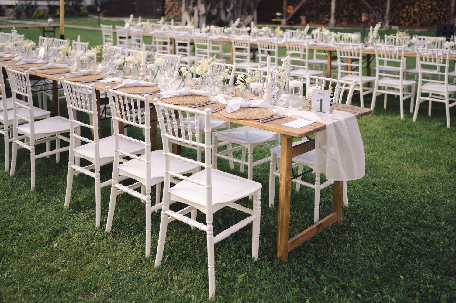 Long rows of elegant white chairs and rustic wooden tables set up on a lawn for an outdoor wedding reception.
