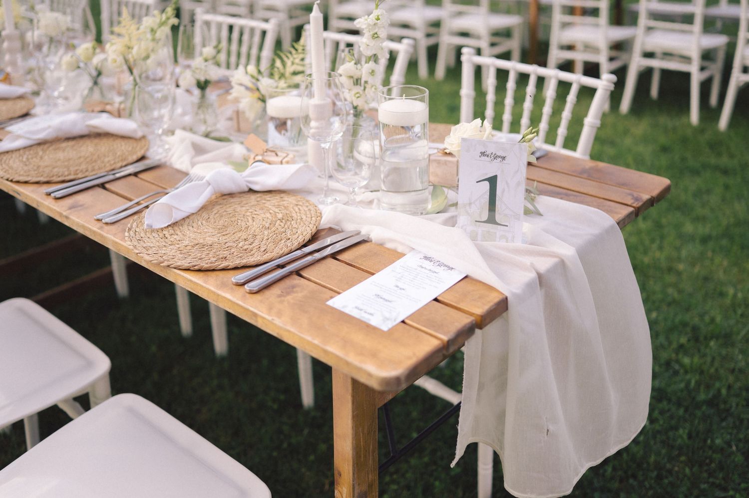 Elegant outdoor wedding table setting with white linen runner, woven placemats and white chiavari chairs on grass.