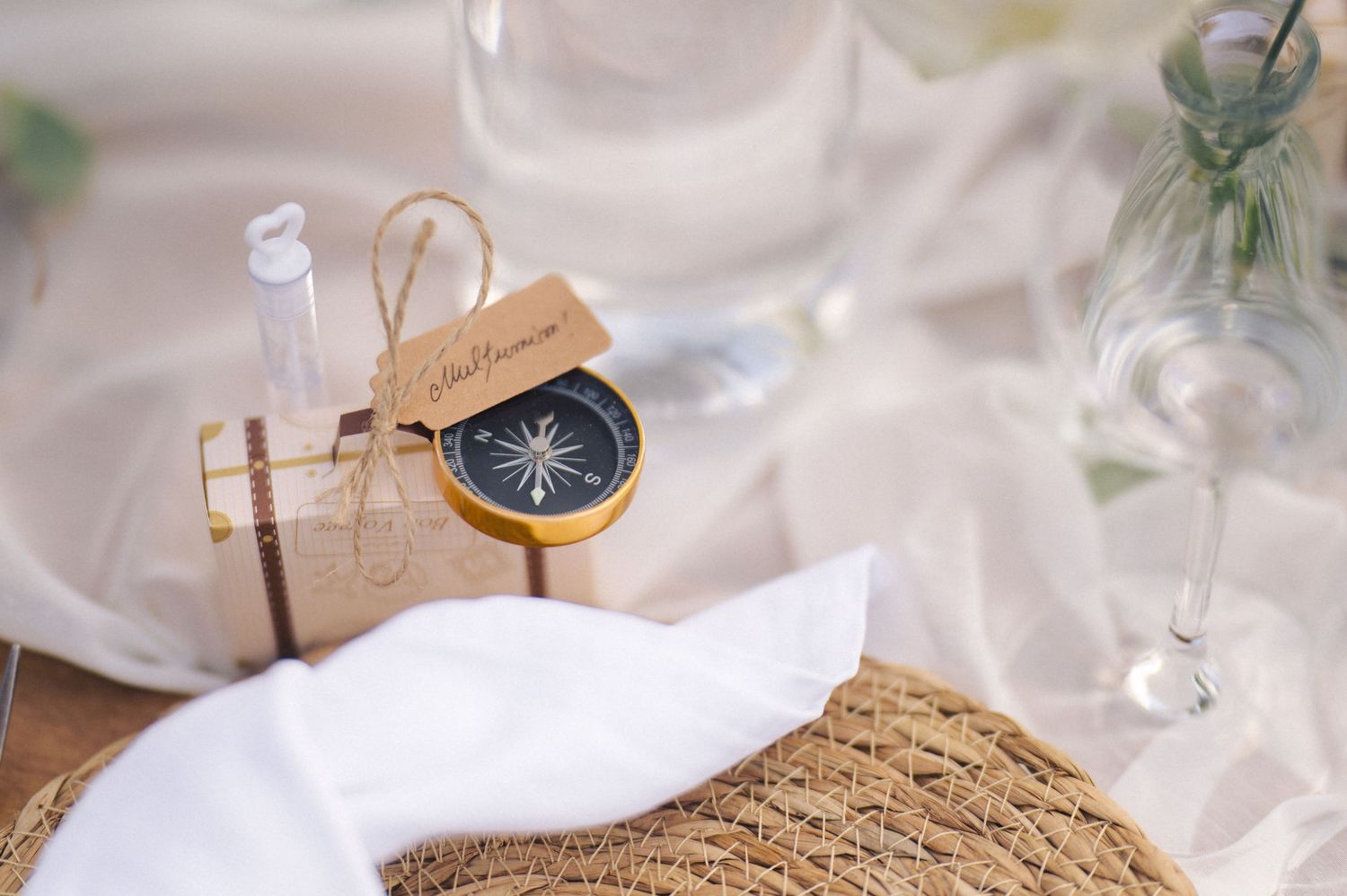 Vintage compass and white napkin on rustic woven placemat with glass decor elements in soft lighting.