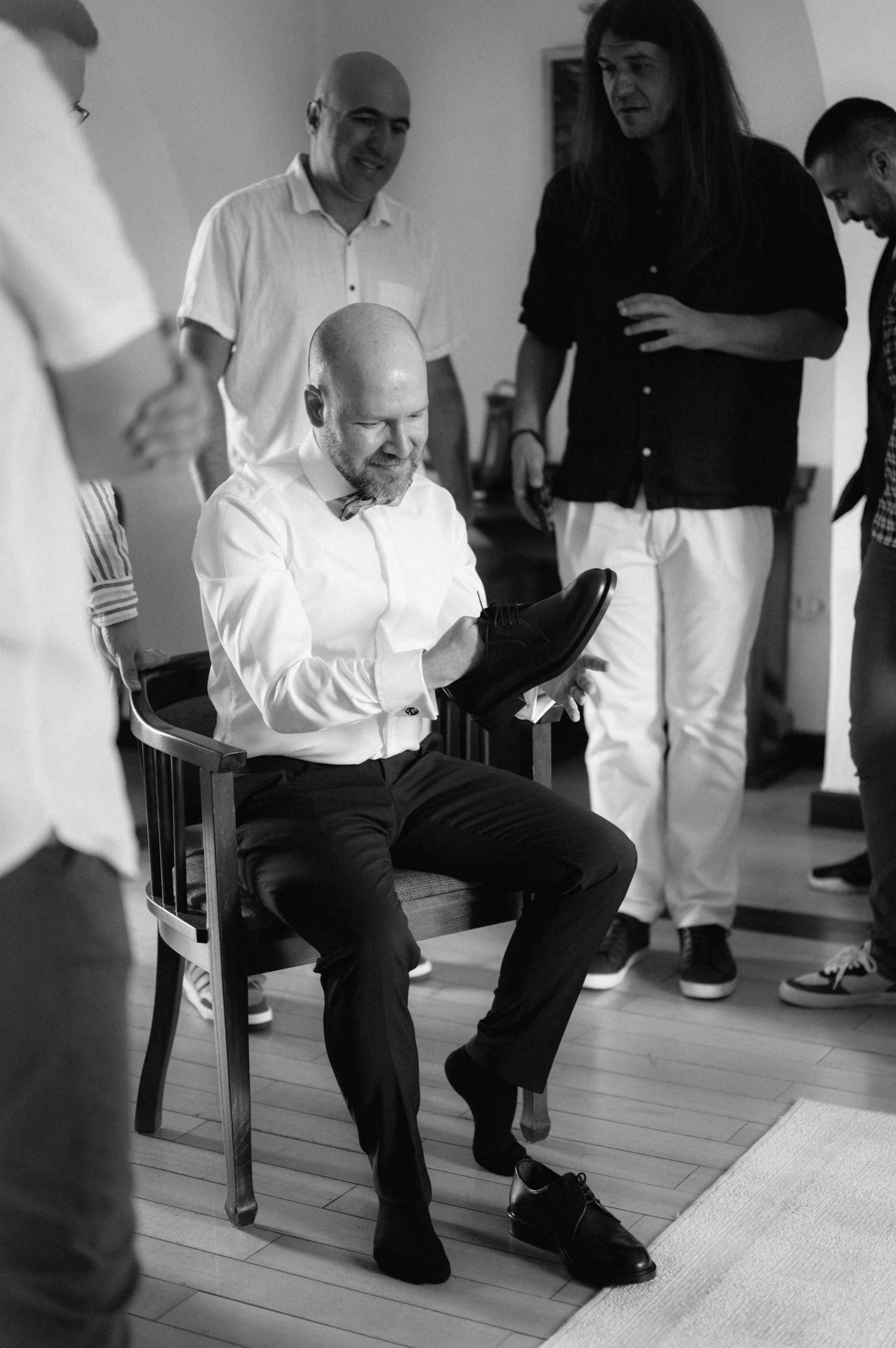 A person sits in a wooden chair looking down at something while others stand around in a bright room with wood flooring.