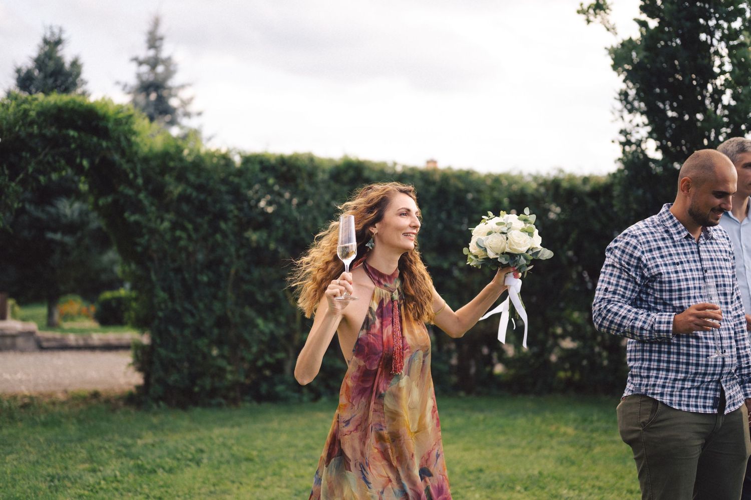 People celebrate outdoors on a lawn with one person holding a white floral bouquet while another stands nearby.