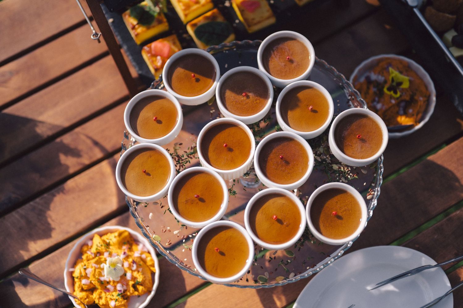 Small cups of butternut squash soup arranged in rows on a wooden surface with garnish and golden lighting.