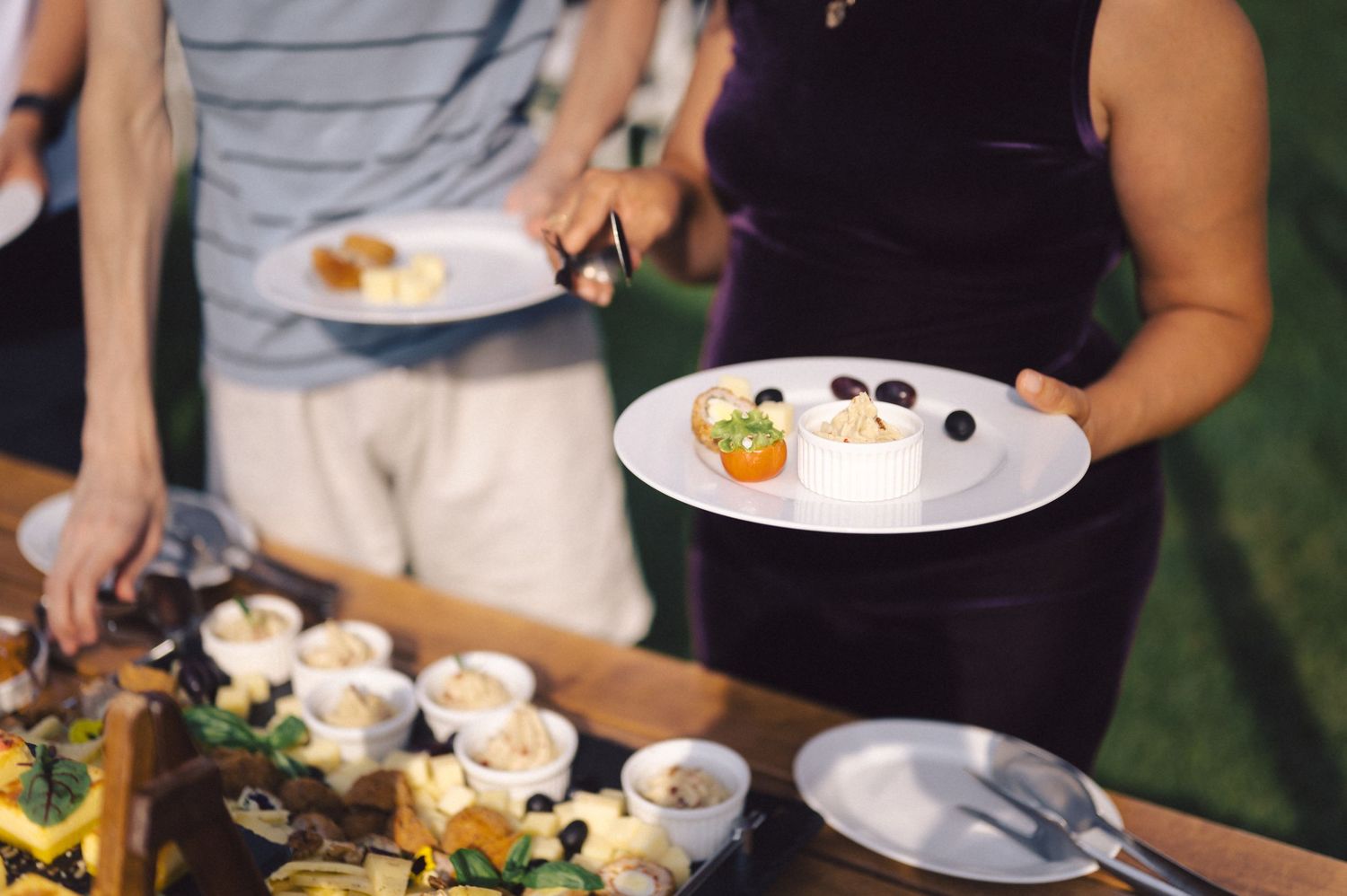 People serving themselves from a buffet table with plates of food and appetizers at an outdoor event.