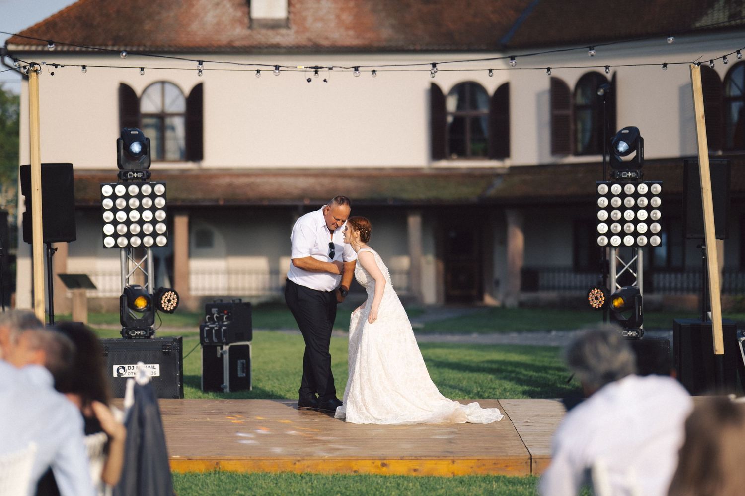 A couple shares their first dance on an outdoor stage at sunset with spotlights and rustic venue backdrop.