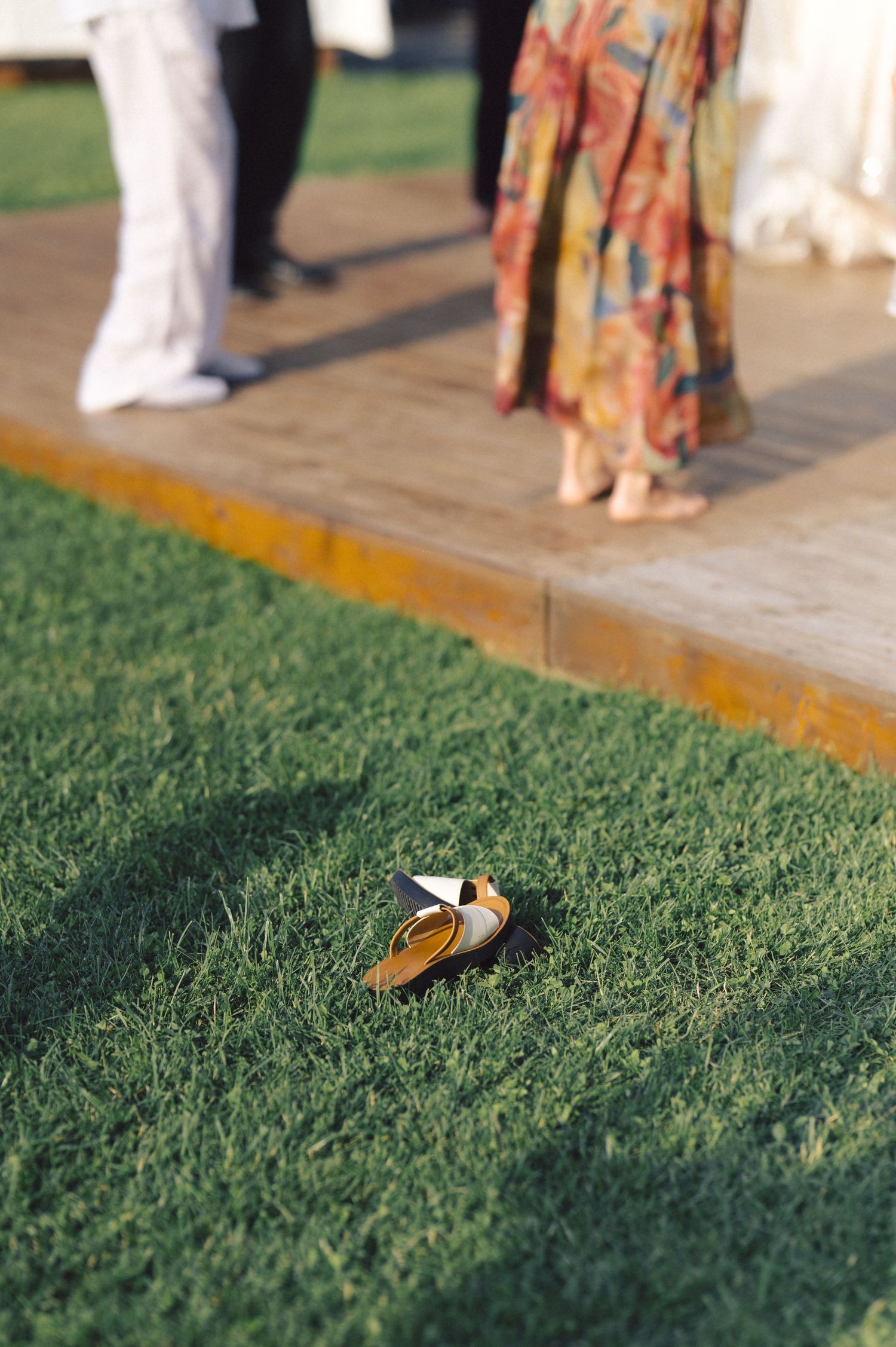 A pair of women's shoes lies on green grass next to a wooden deck where people in formal attire are standing.