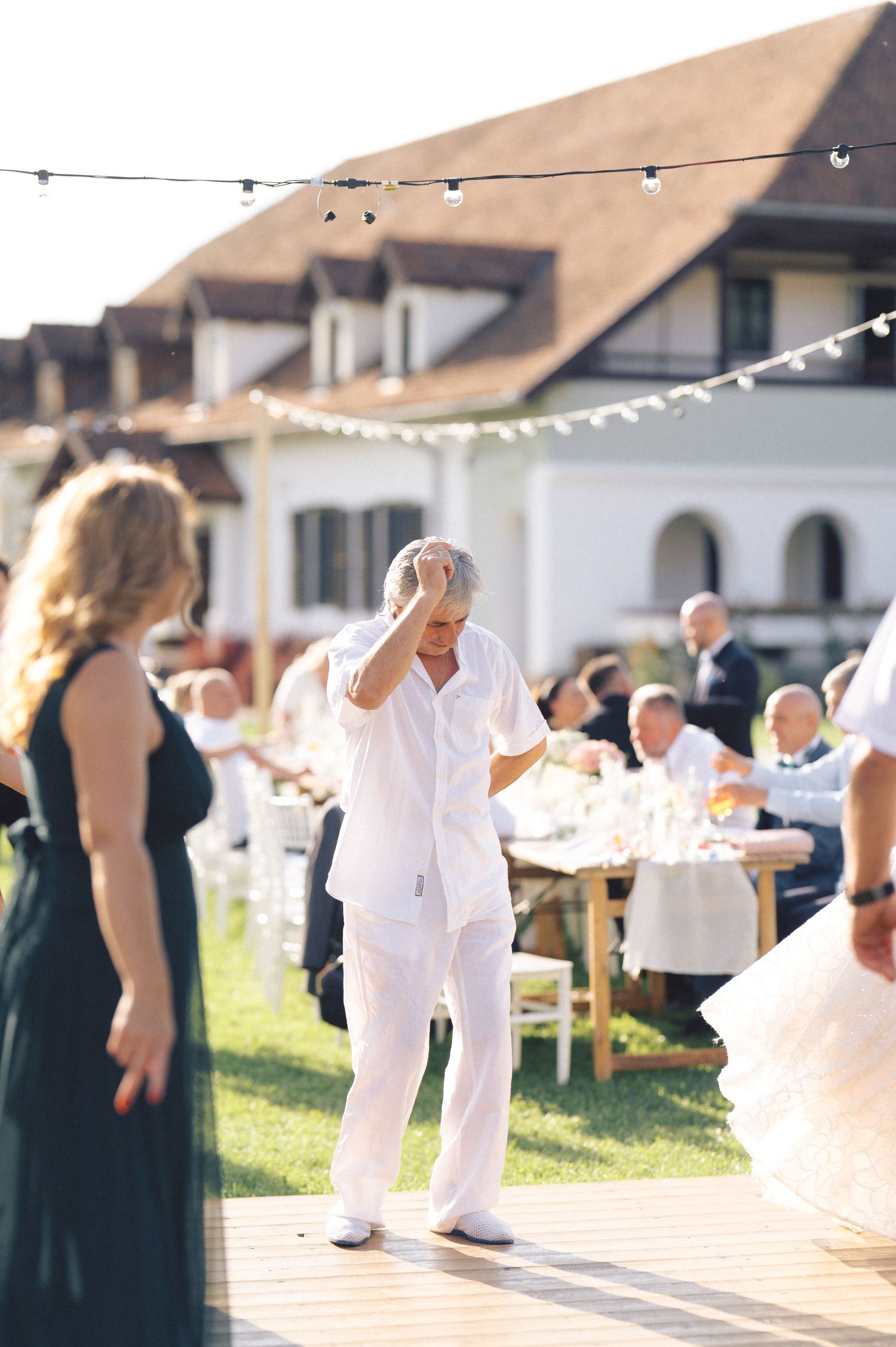 People celebrating at an outdoor party with string lights and wooden tables near a white building in warm sunlight.