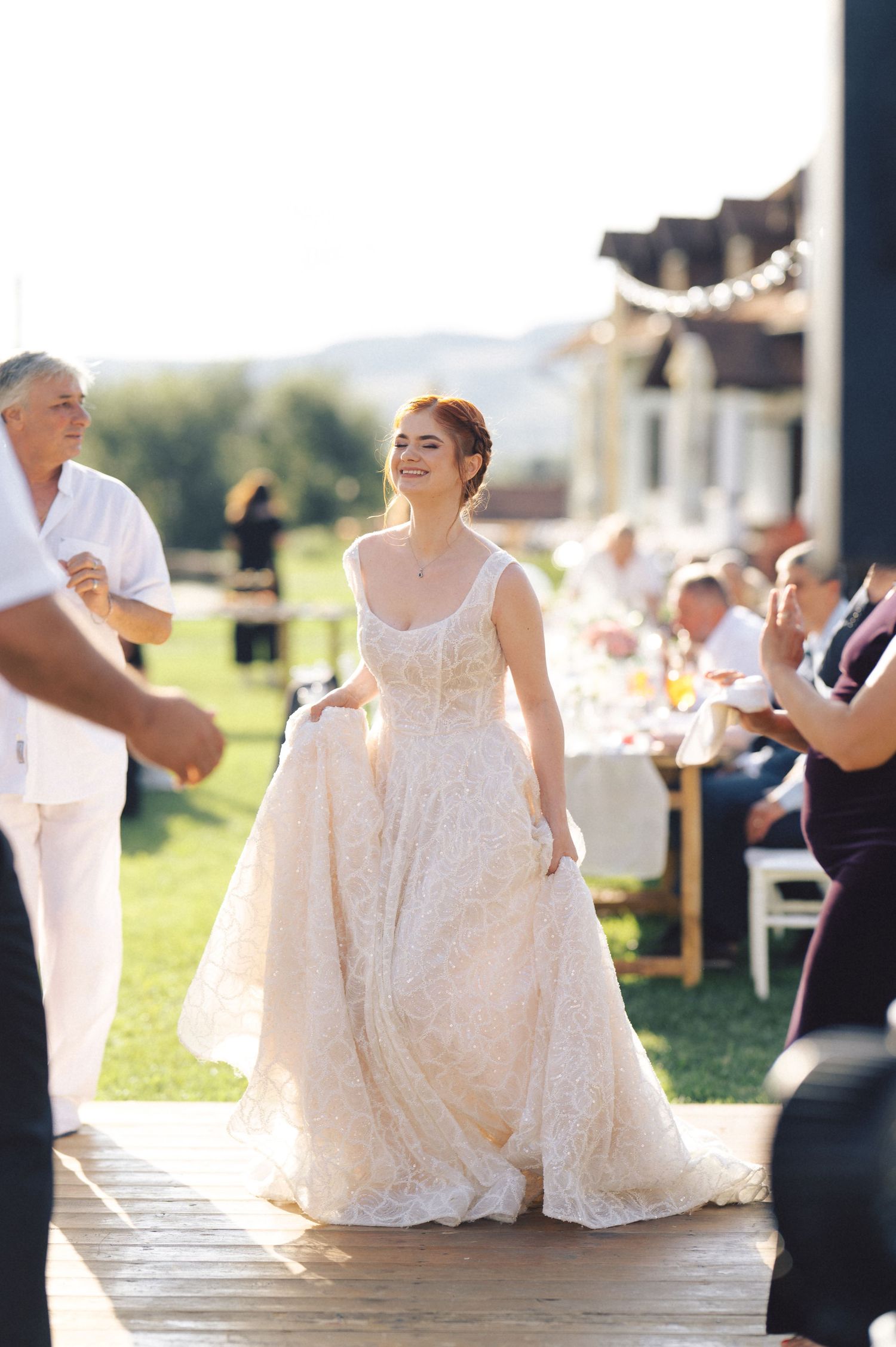A bride in a flowing white lace gown smiles during an outdoor wedding reception on a sunny day.