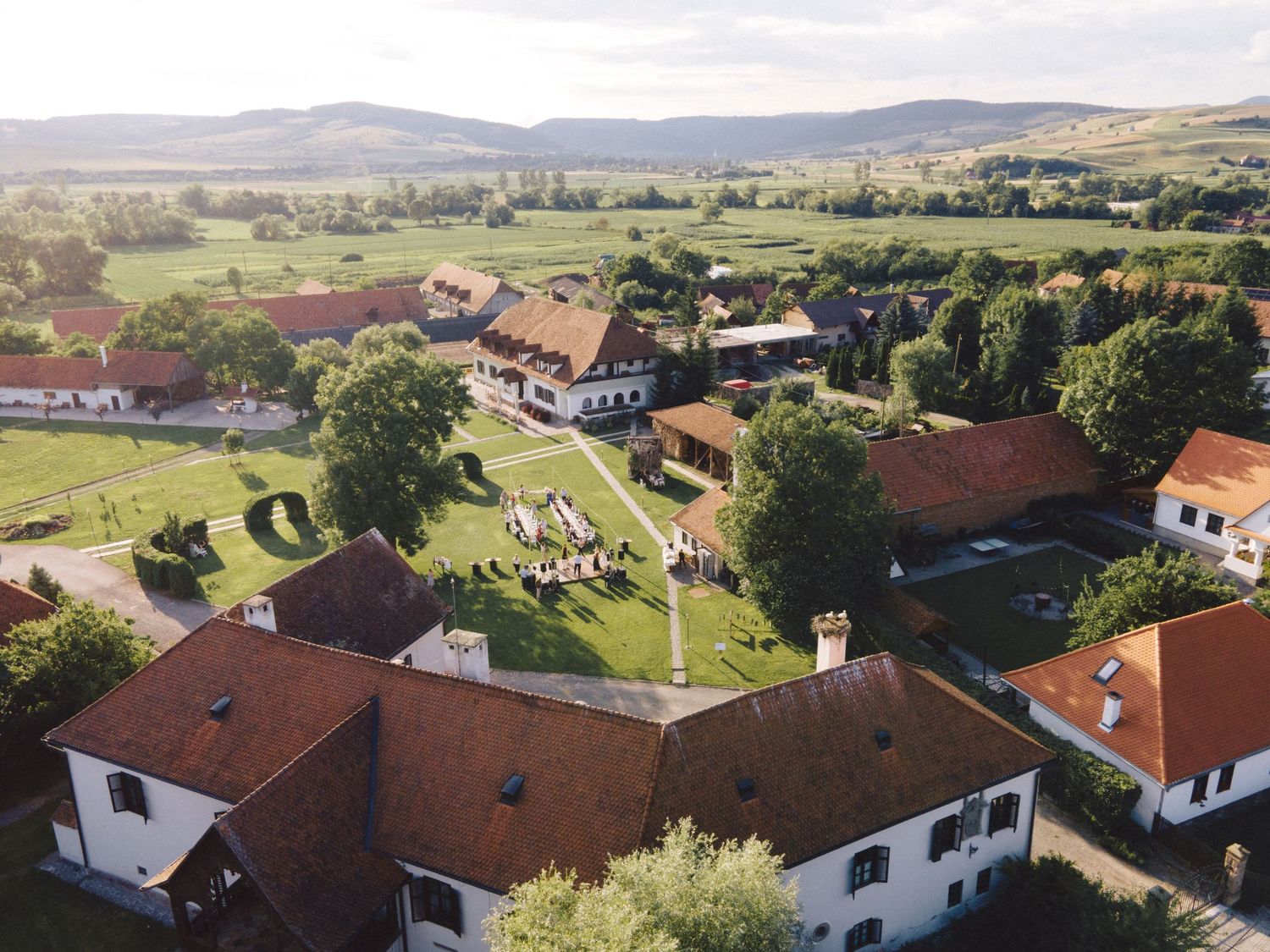 Aerial panoramic view of a rural village with red-roofed houses nestled among green fields and rolling hills.
