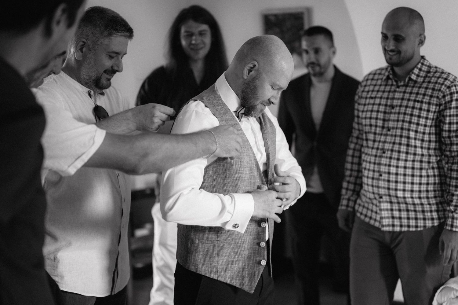 Black and white photo of groomsmen helping adjust a vest and formal attire while getting ready before a wedding ceremony.