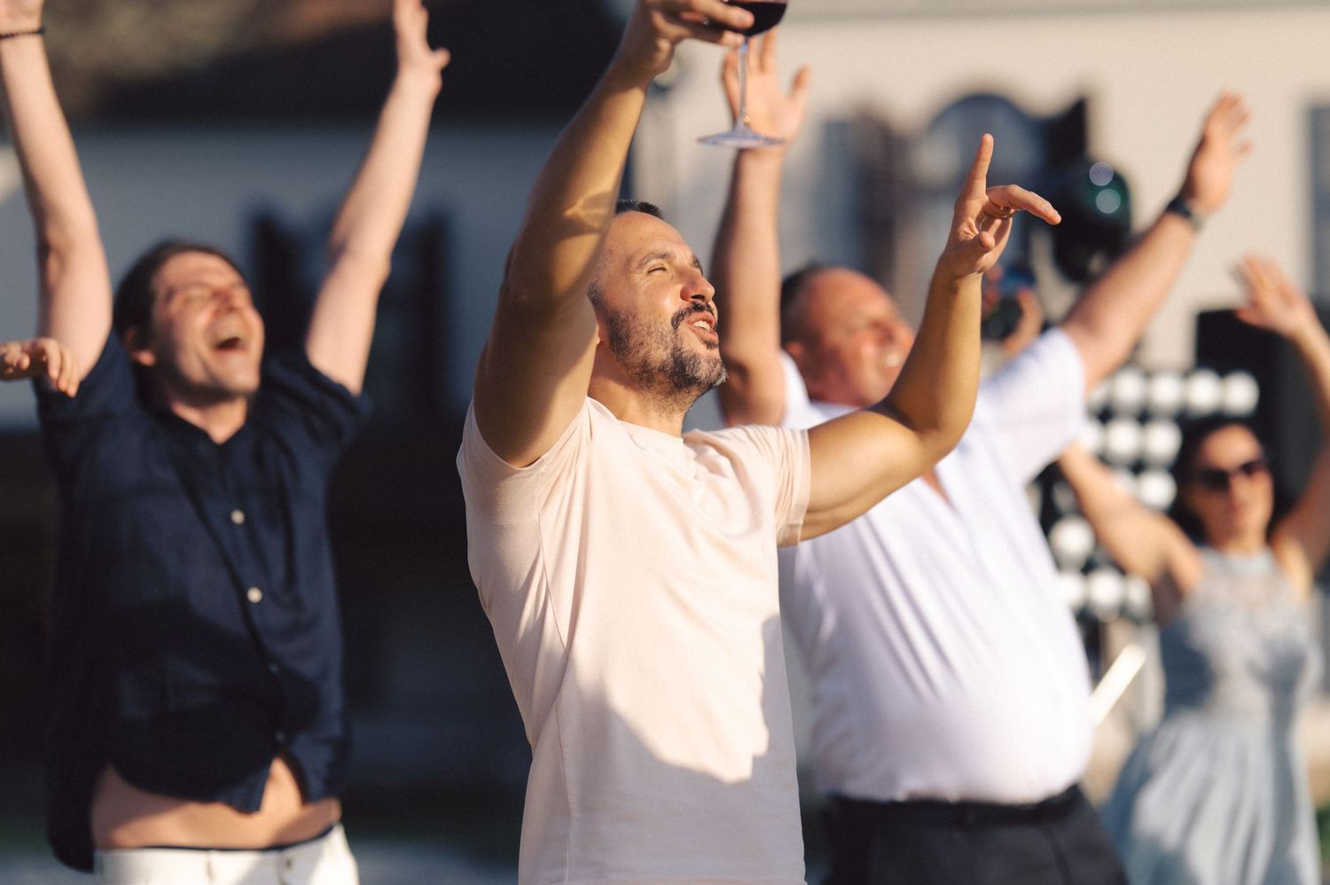 A group of spectators cheer excitedly with raised arms while enjoying an outdoor event on a sunny day.