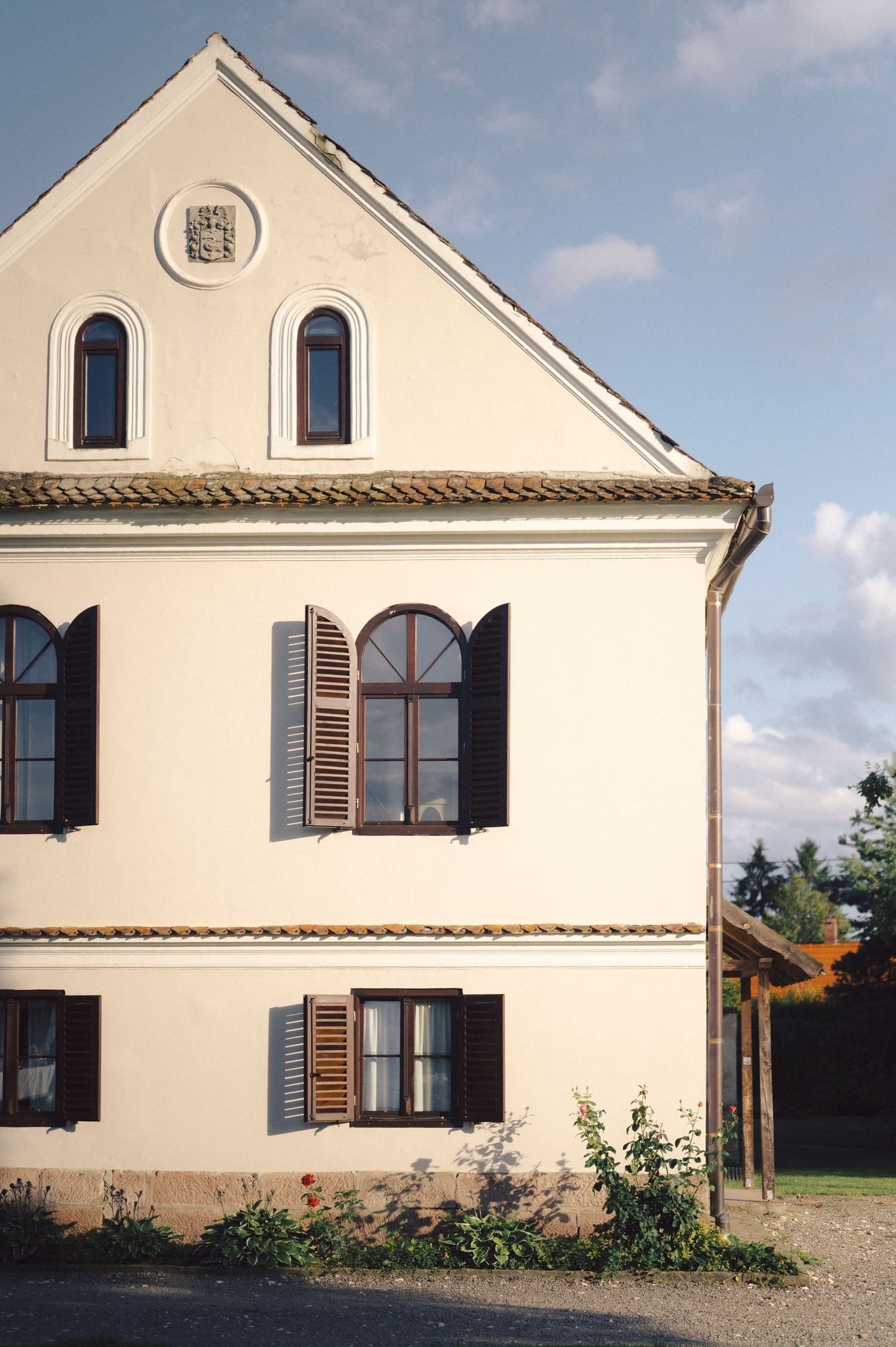 White historic house with brown shutters, arched windows, and a triangular gable featuring two narrow windows against blue sky.