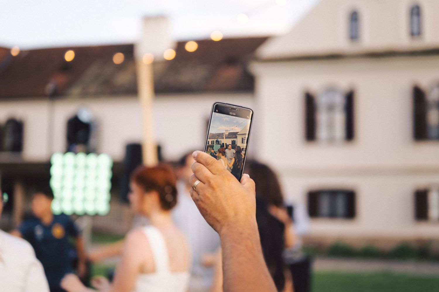 Hand holding smartphone taking photo at outdoor wedding venue with white colonial-style building in background.