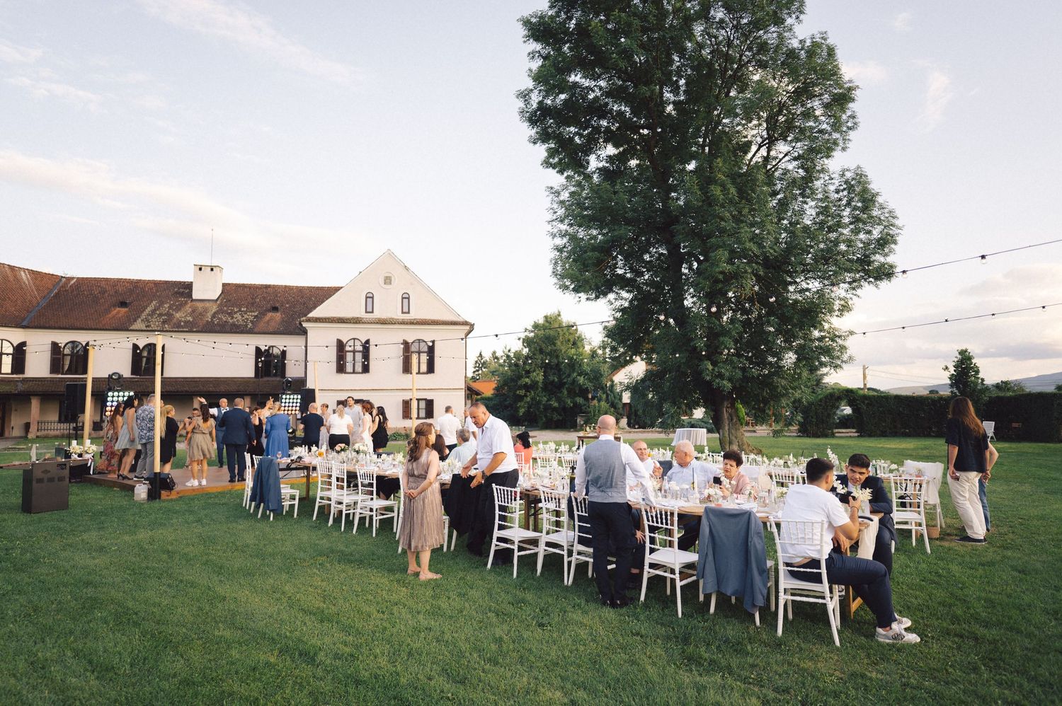 People seated at long outdoor tables for a dinner gathering on a lawn in front of a white country house.