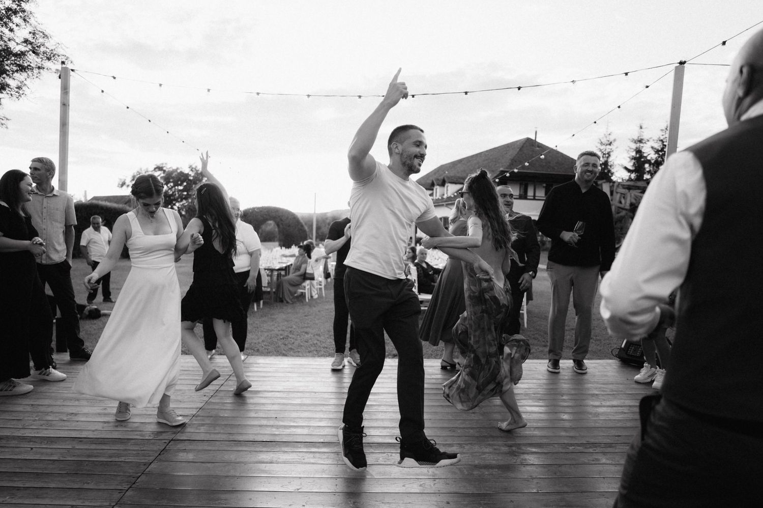 Black and white photo of people dancing at an outdoor celebration party on a wooden deck with string lights overhead.