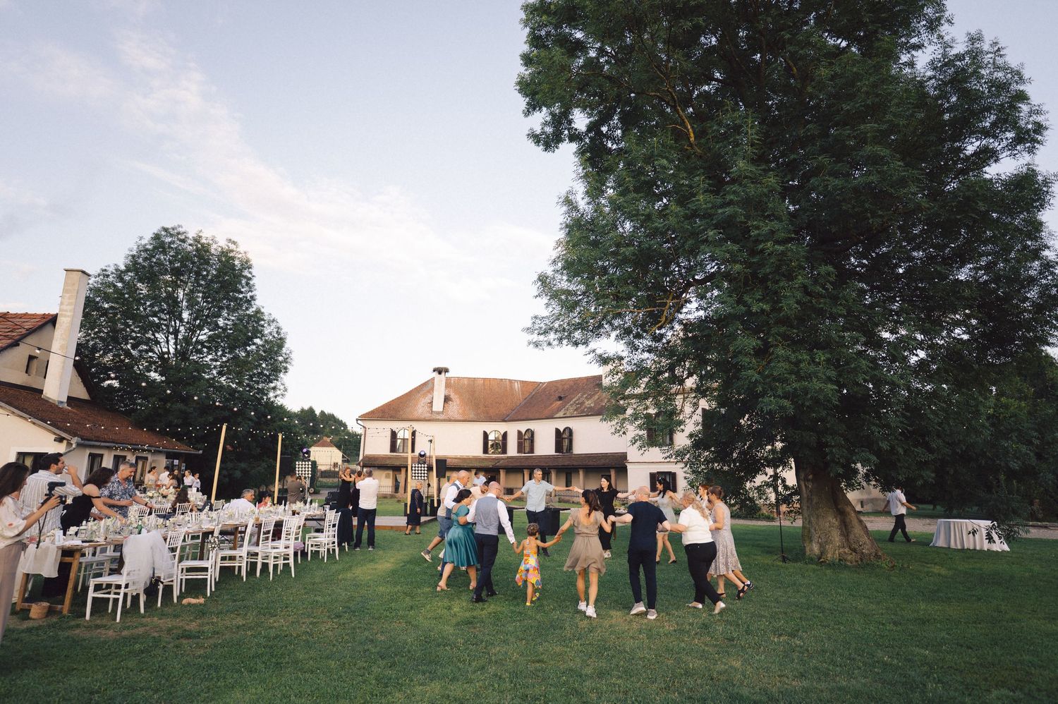 Wedding guests gather under shady trees on a green lawn during an outdoor summer reception celebration.