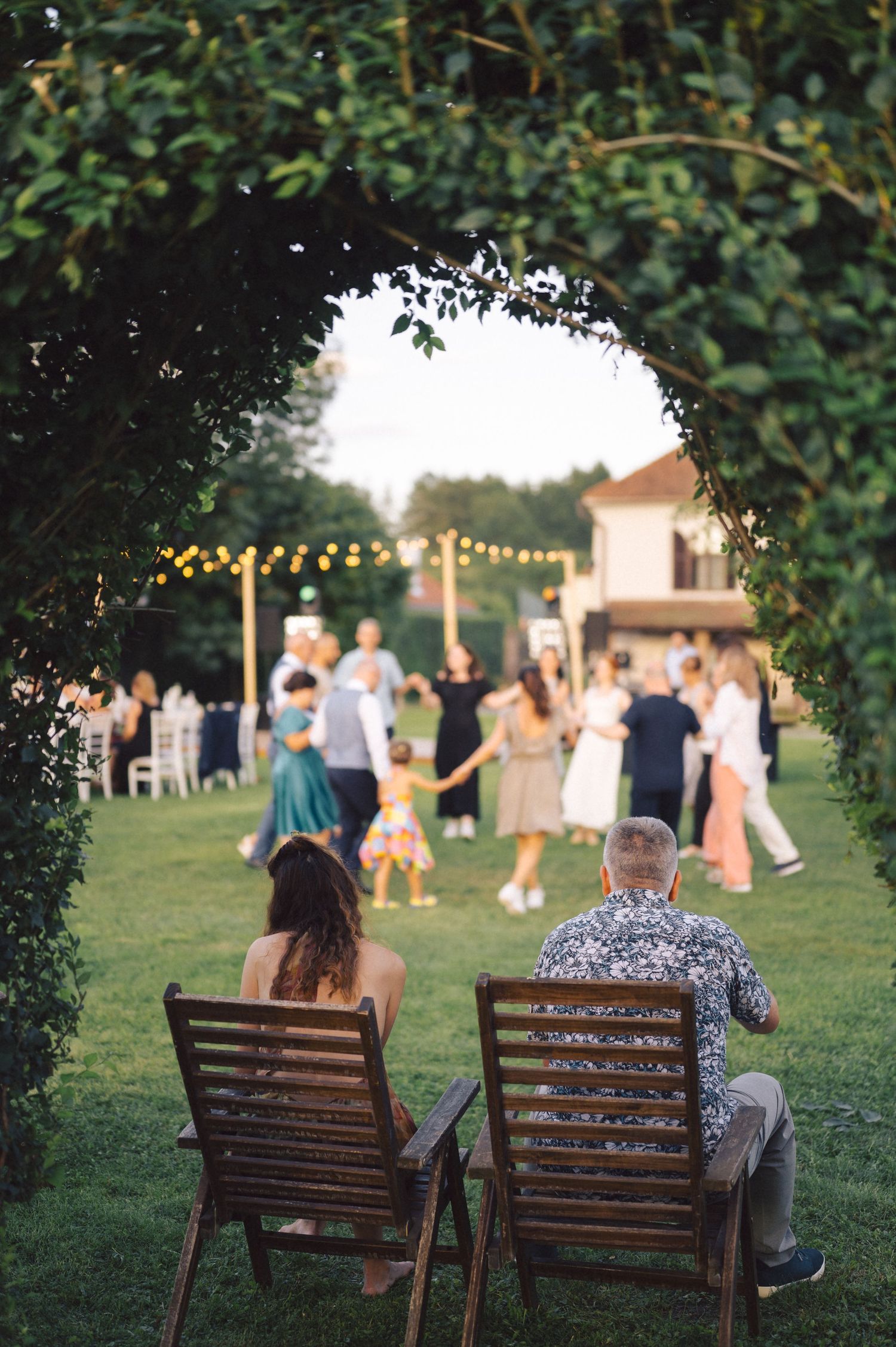 Couple sits on wooden benches watching guests dance at an outdoor wedding reception with string lights at dusk.