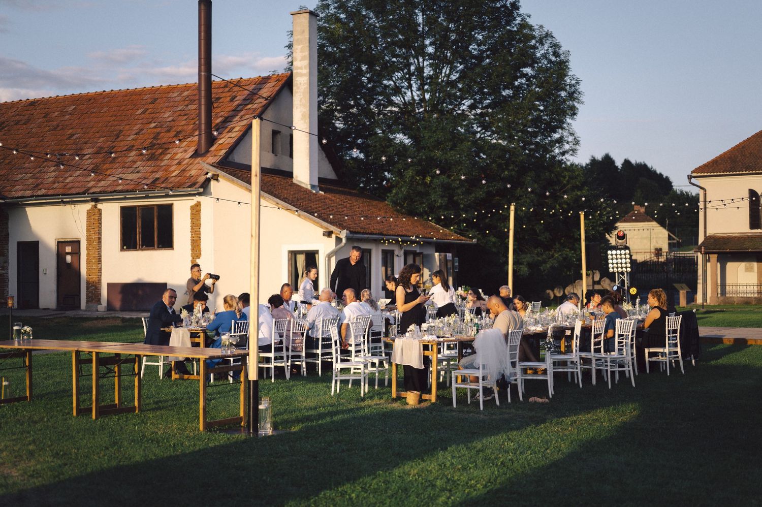 Outdoor evening gathering with people seated at white tables on a lawn next to a yellow house with terracotta roof.