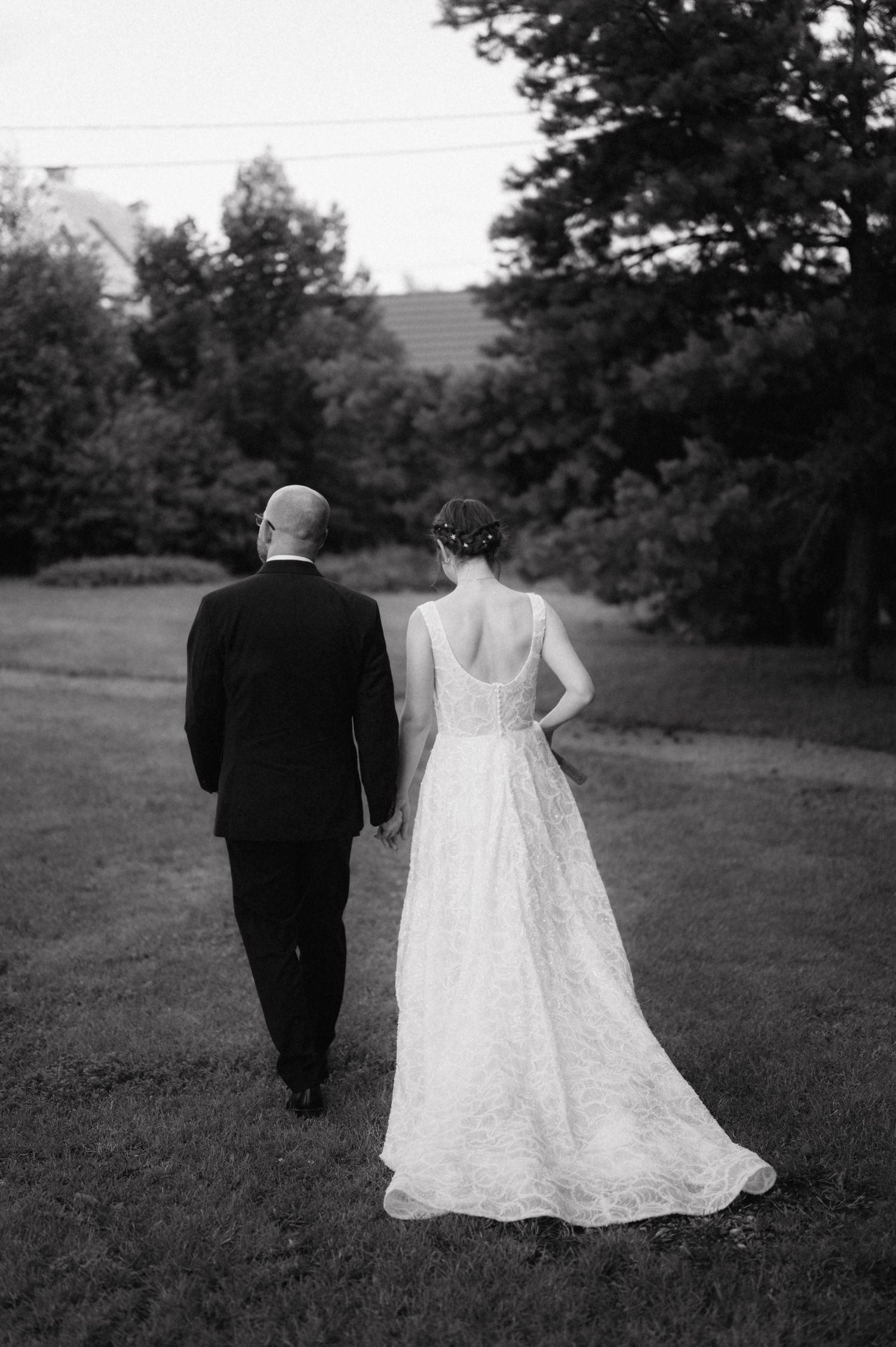 Black and white photo of couple walking away on grass, bride wearing lace wedding dress with low back detail.