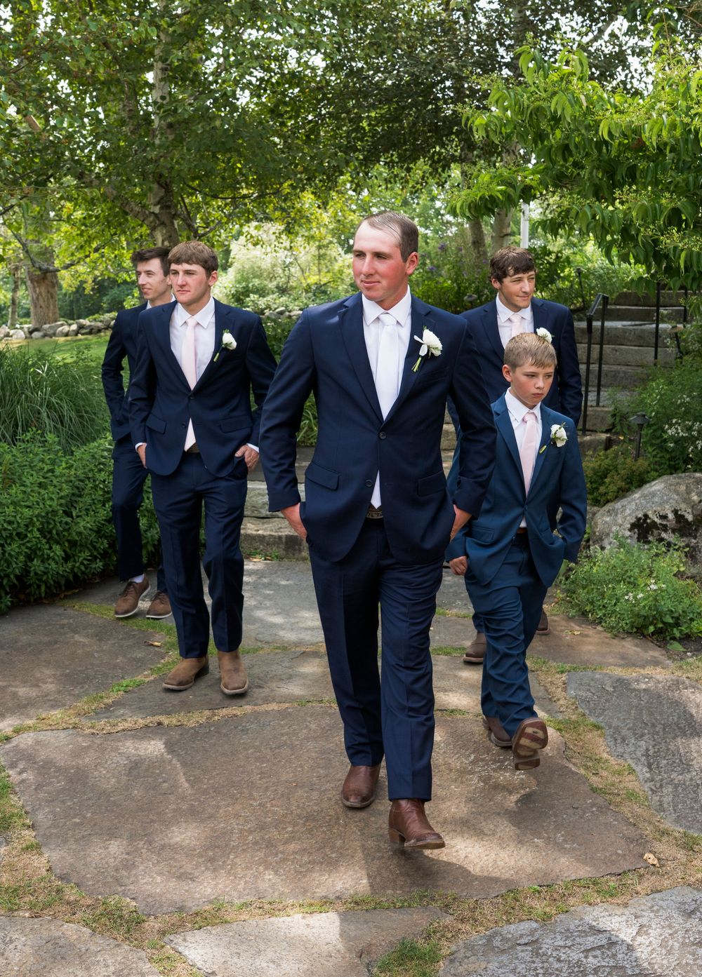 Groomsmen in navy suits walk along garden path before wedding ceremony.