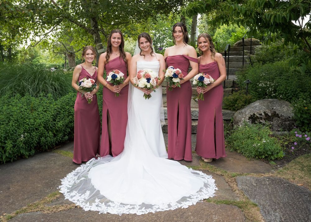 Bridal party in dusty rose dresses poses outdoors with trailing white wedding gown.